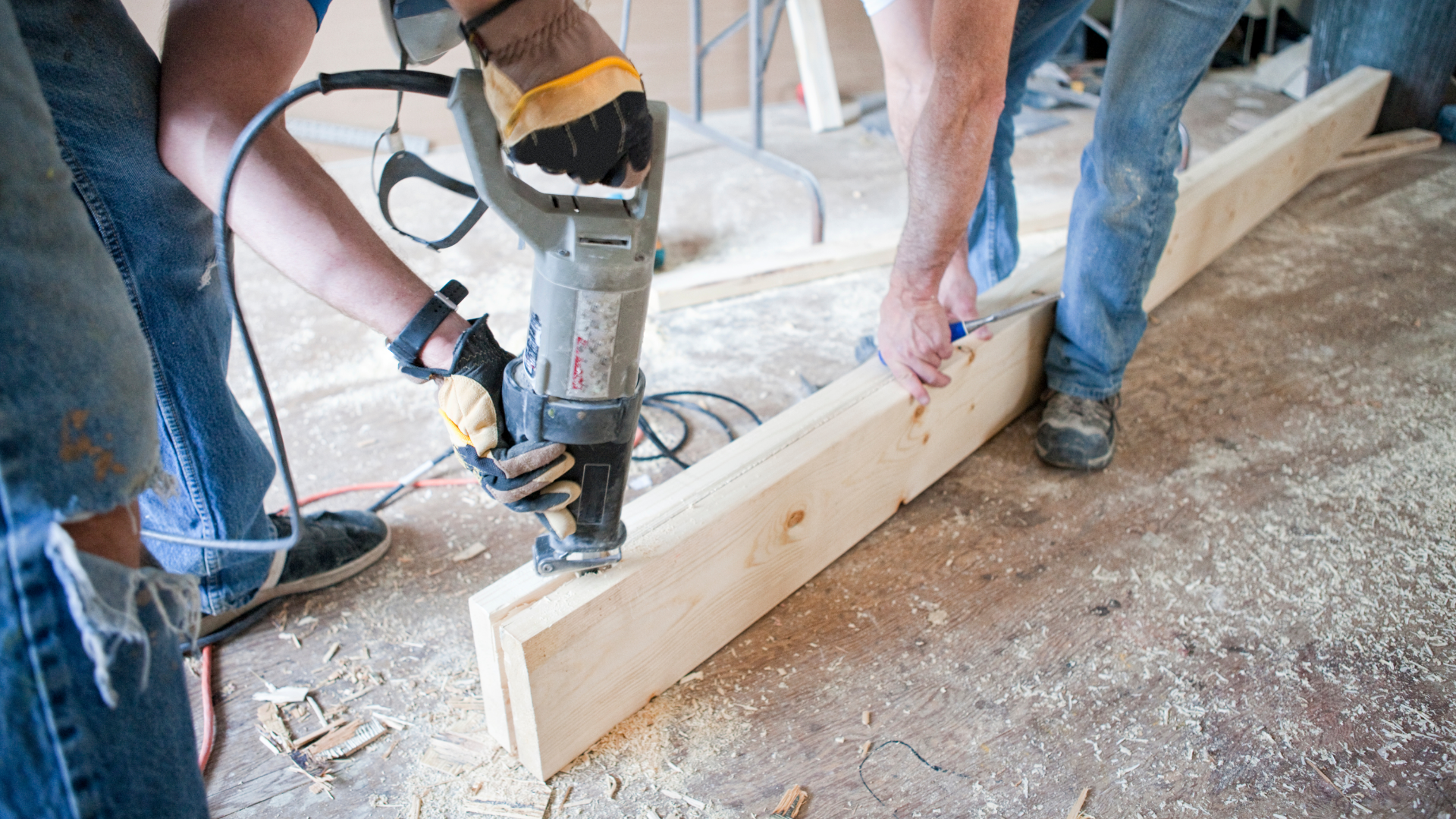 Two people working with a power tool on a wooden beam in a room under construction.