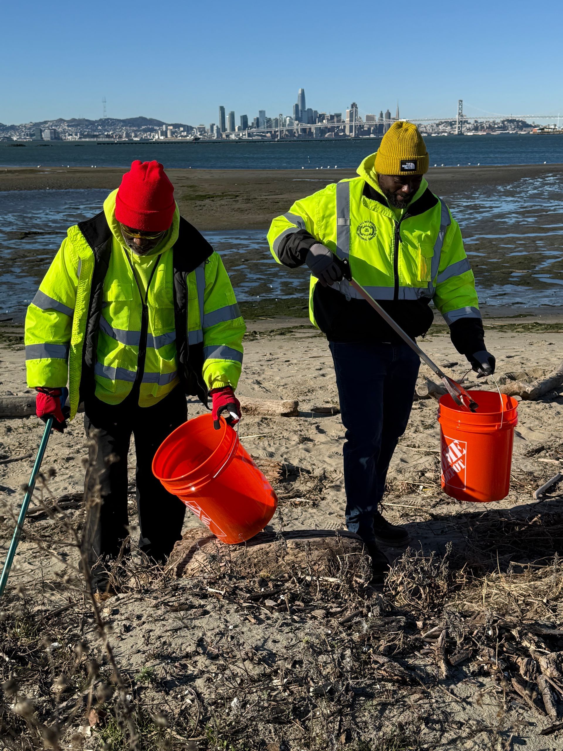 Two people in bright vests clean up trash on a beach, San Francisco skyline in the background.