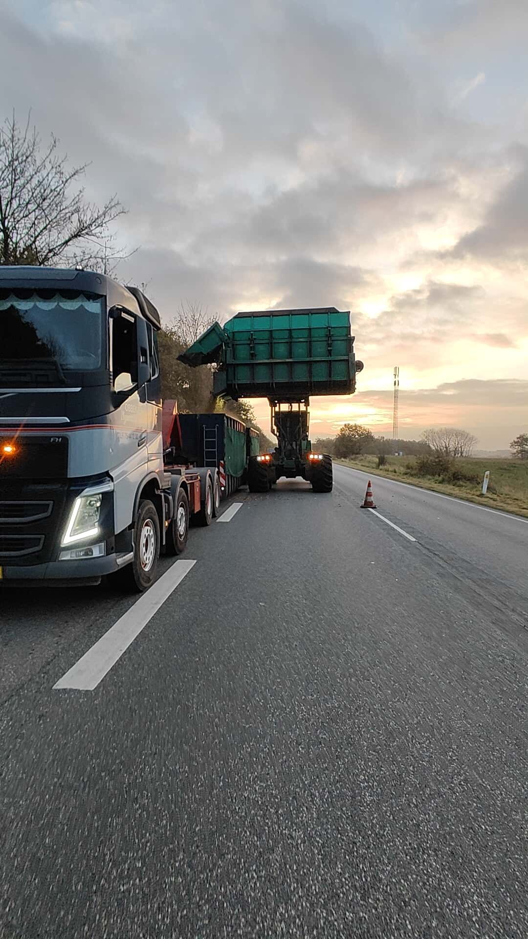 To lastbiler holdt stille på en motorvejsskulder ved solnedgang, den ene med en stor grøn last.