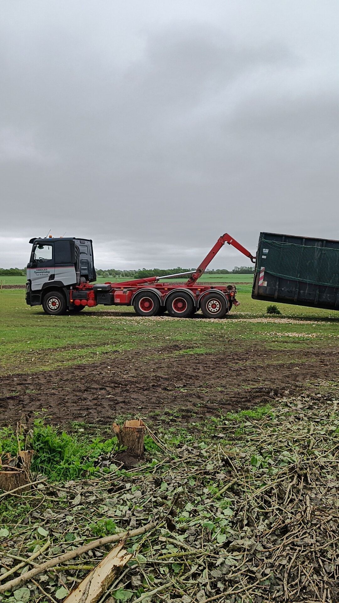 Traktor med rød læssearm i en mudret mark løfter en tippet grøn trailer under overskyet himmel