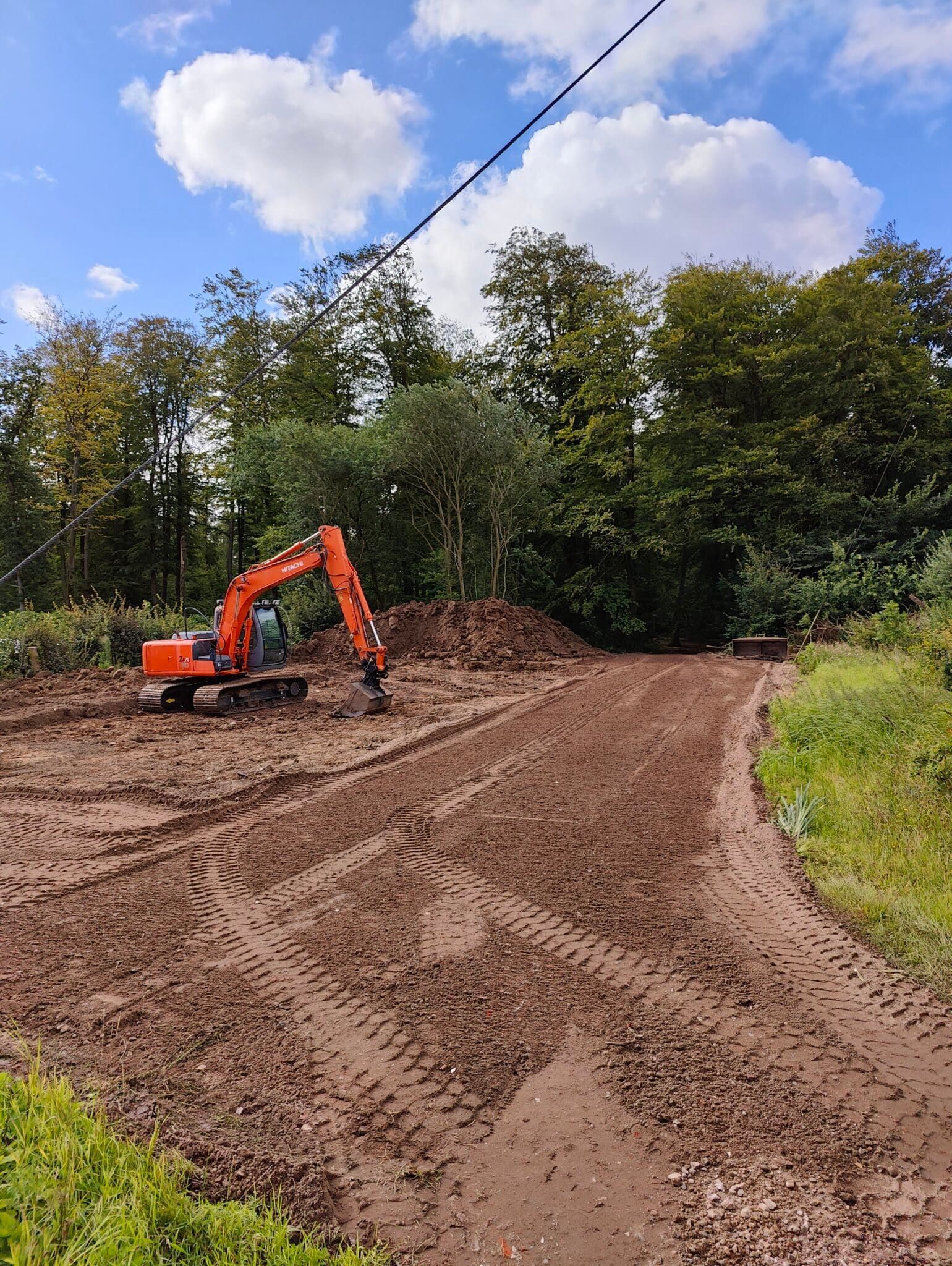 Orange gravemaskine på en mudret grusvej ved siden af en skov under en overskyet blå himmel