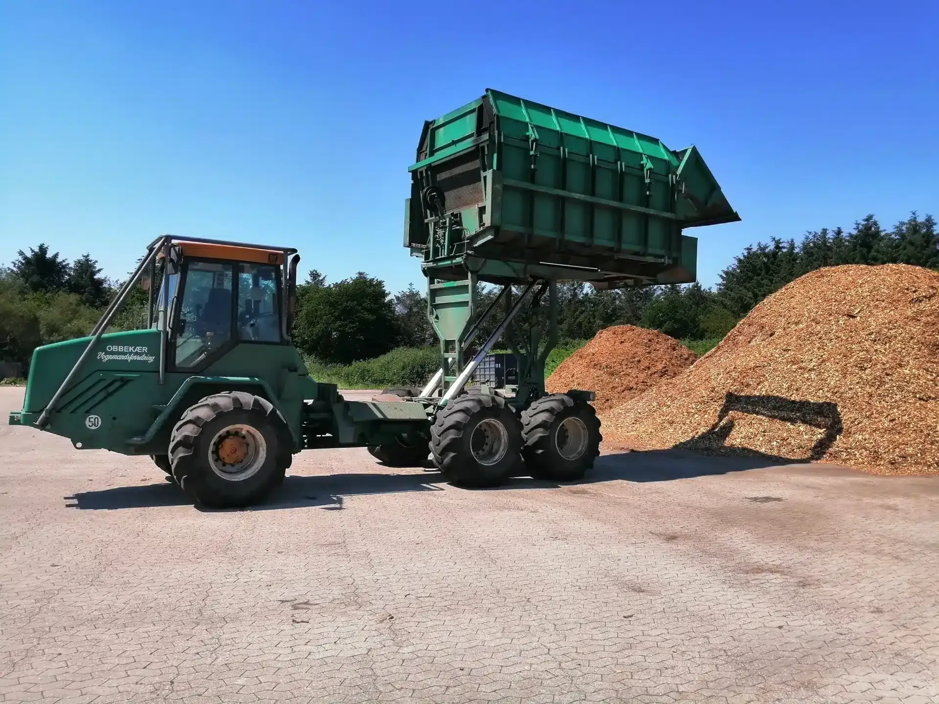 Grøn traktor tipper en masse træflis i en bunke på en gårdsplads langs vejen.