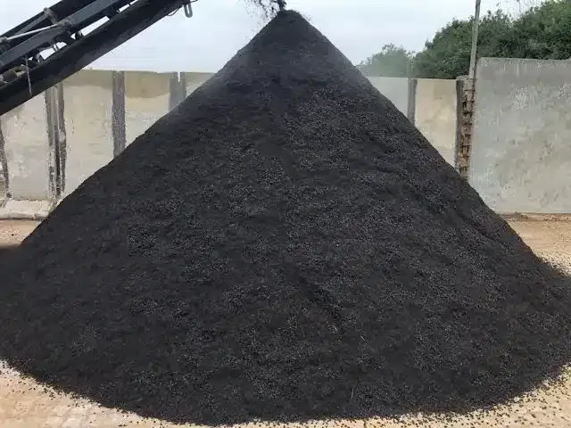 Stor sort grusbunke på en udendørs industrigrund med et transportbånd over og betonvægge bagved.