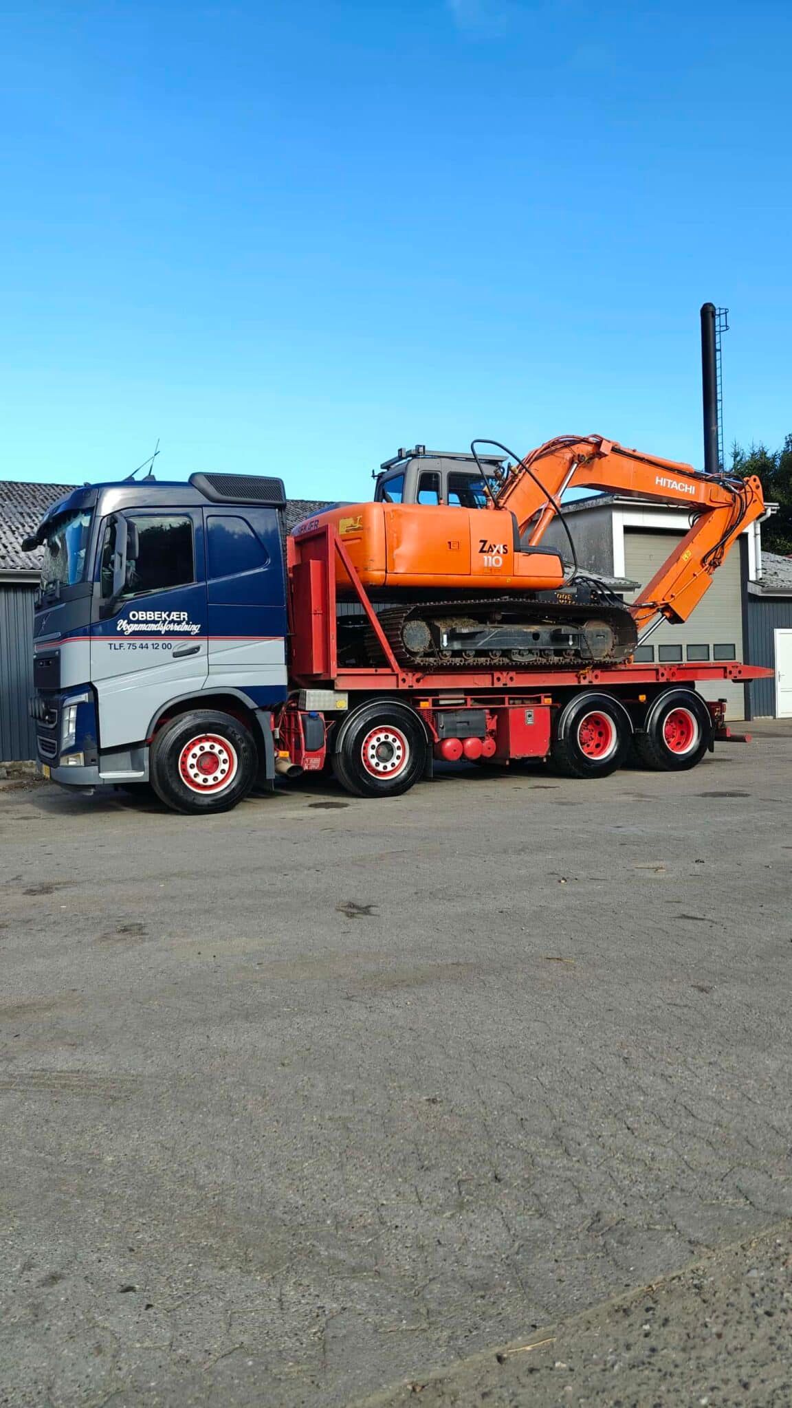 Orange gravemaskine på en rød lastbiltrailer parkeret på en åben plads under en blå himmel