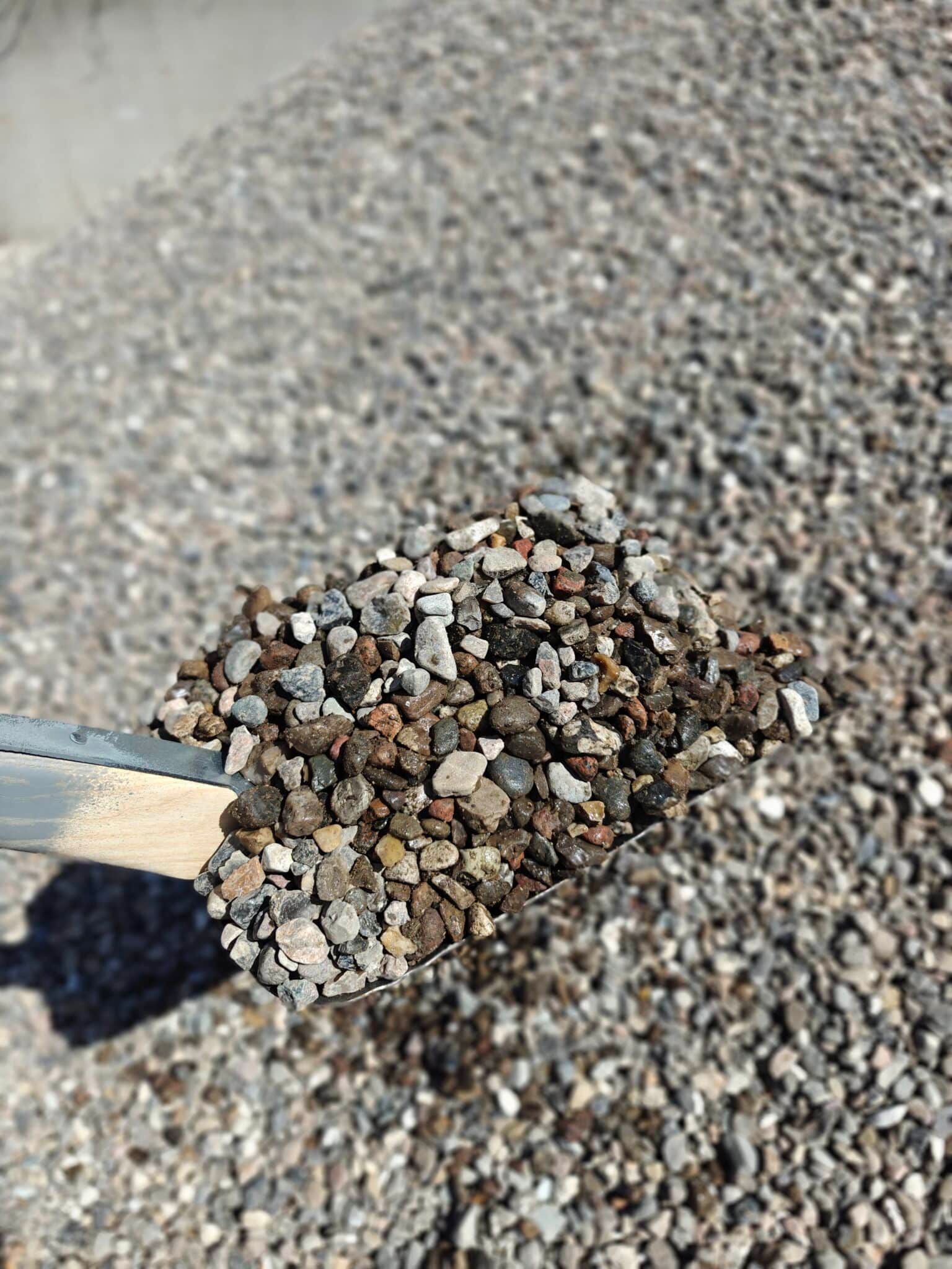 Skovl, der holder en skefuld små grå og brune grus på en grusoverflade.