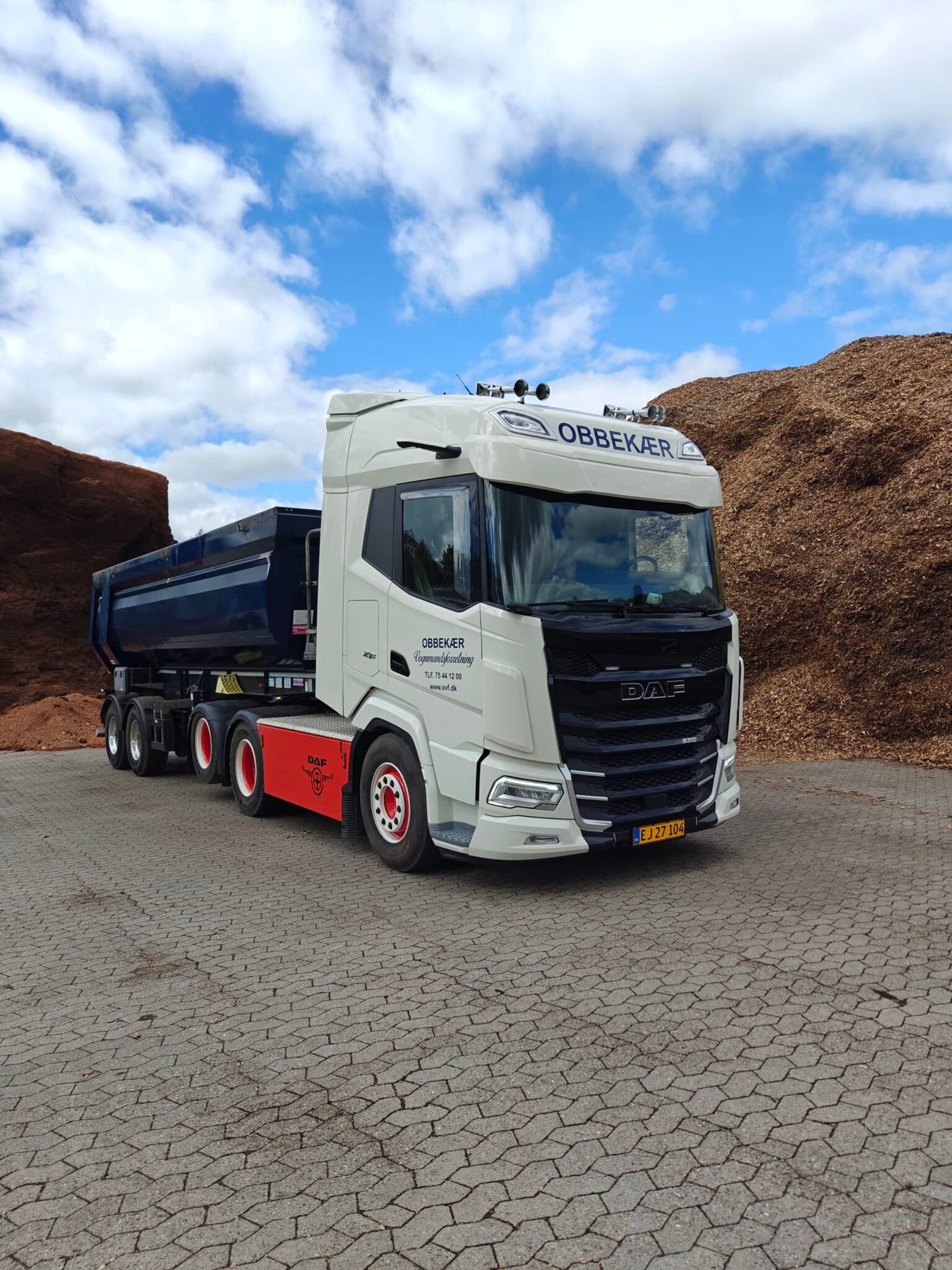Hvid og sort semitrailer parkeret på revnet jord med træflis under en overskyet blå himmel
