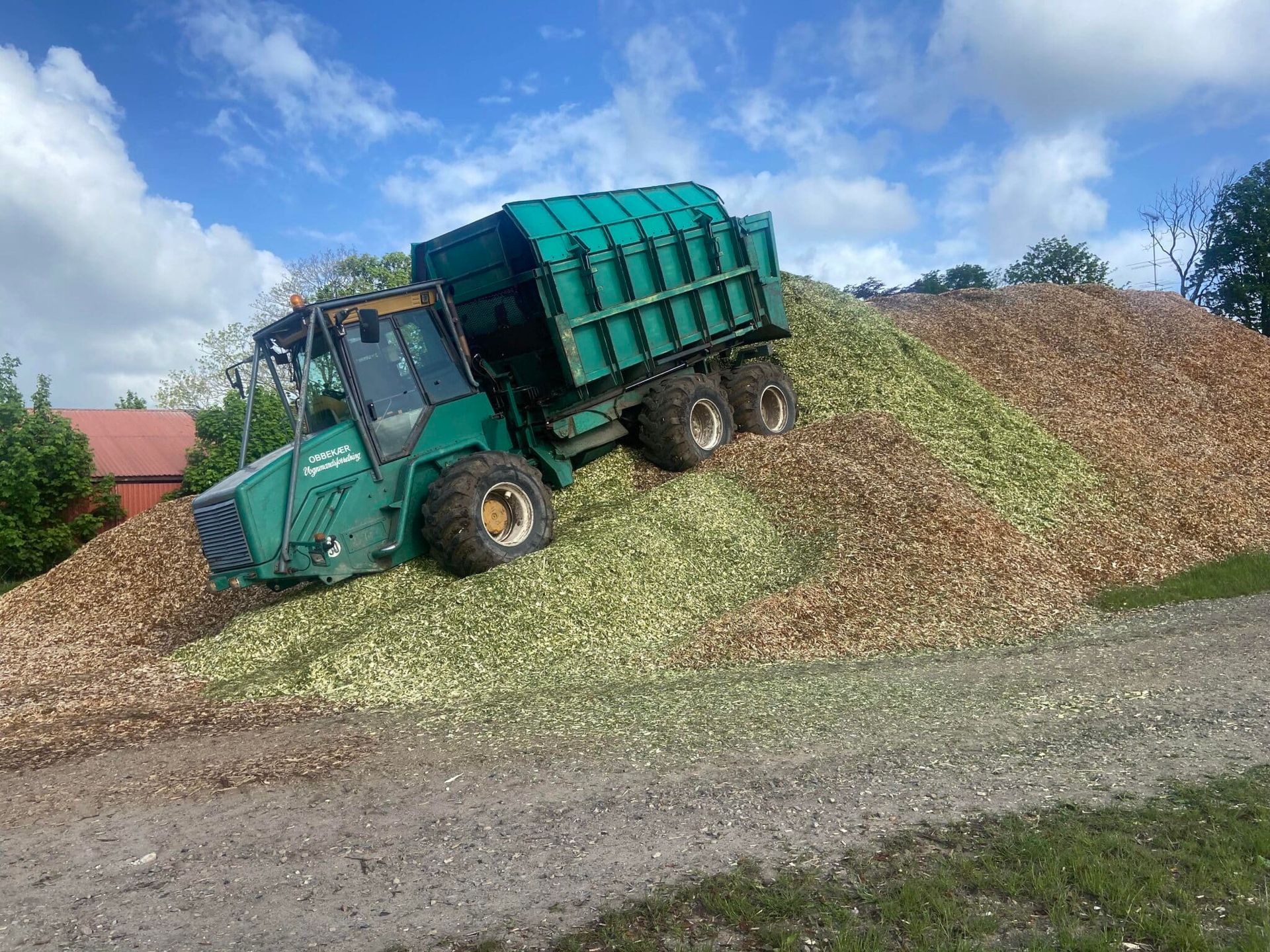 Grøn landbrugsvogn aflæsser hakket ensilage på en stor landbrugsbunke under en overskyet himmel