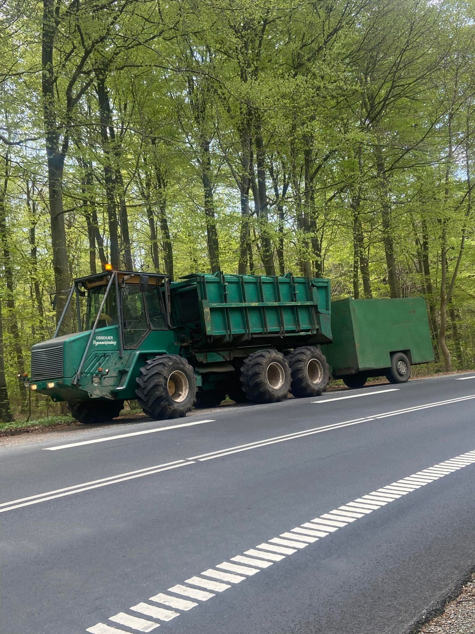 Grøn dumptruck, der trækker en trailer parkeret langs vejkanten ved siden af ​​et skovområde