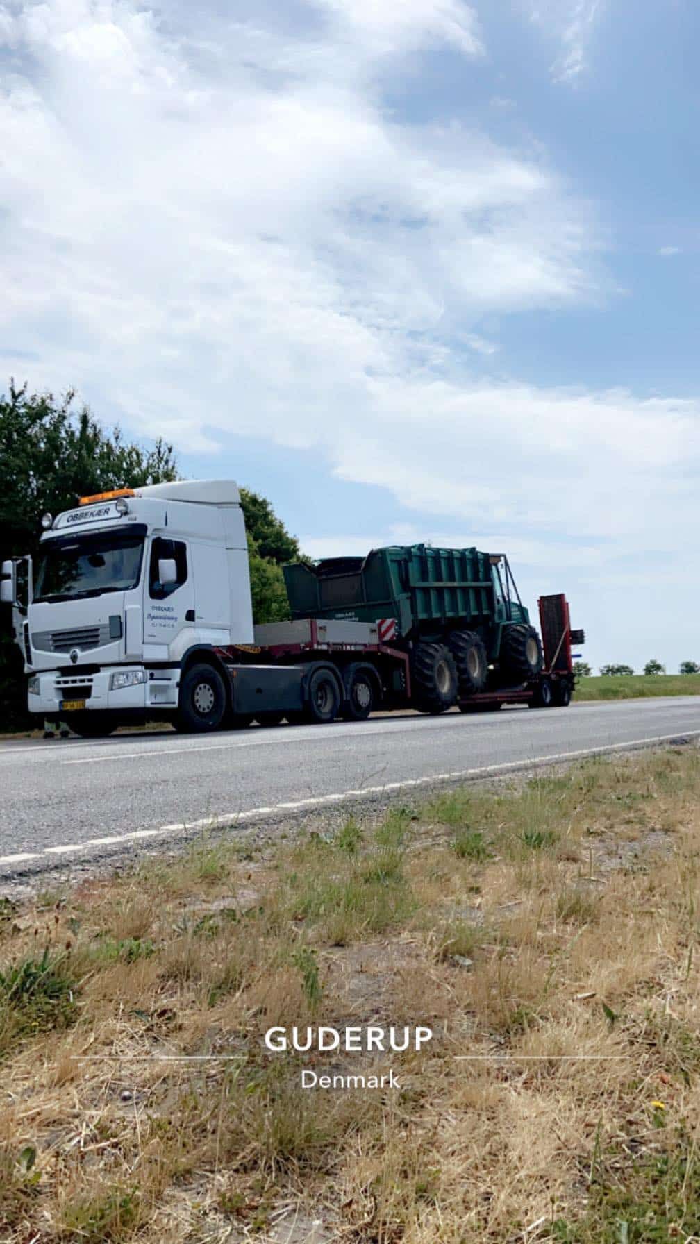 Hvid semitruck, der transporterer en grøn skraldebil langs vejkanten under en overskyet himmel