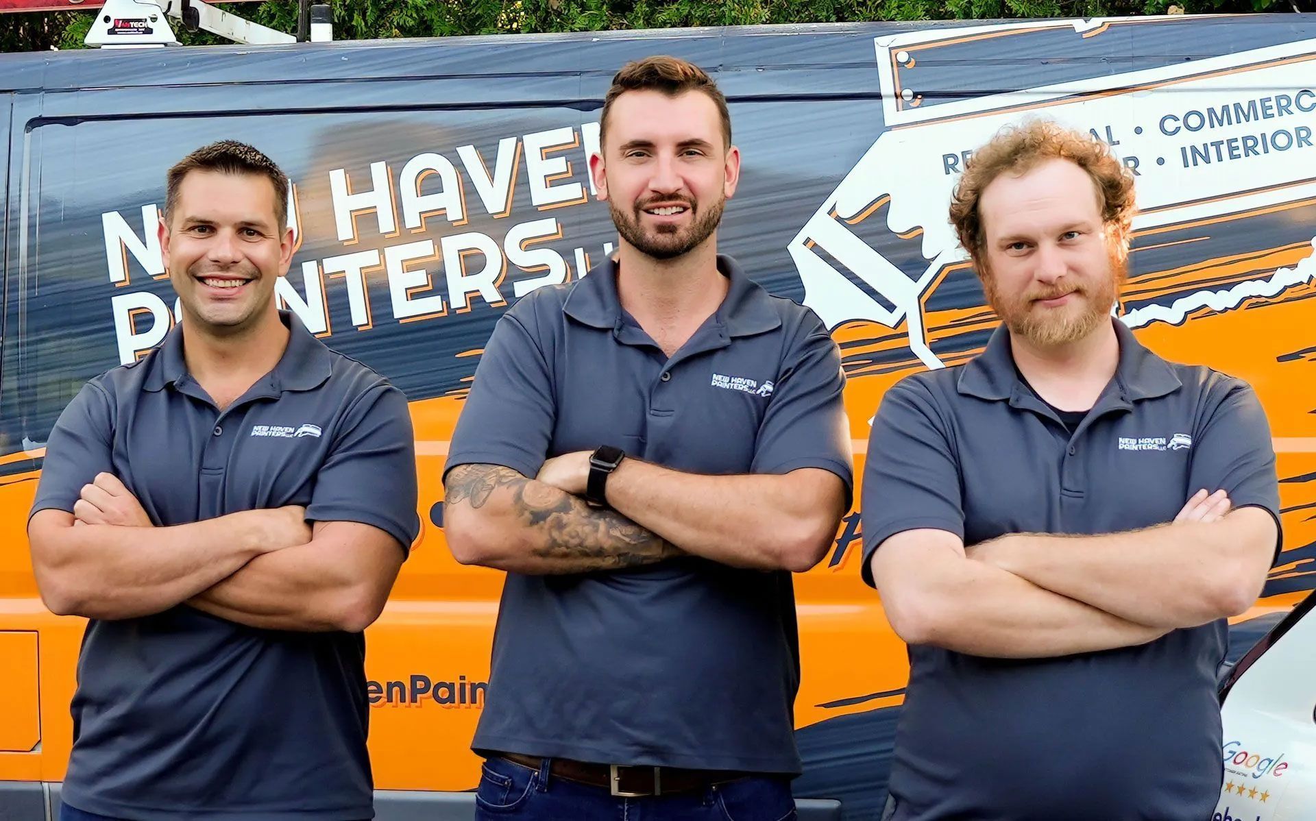 Three men in gray shirts with arms crossed in front of a van. They are smiling.