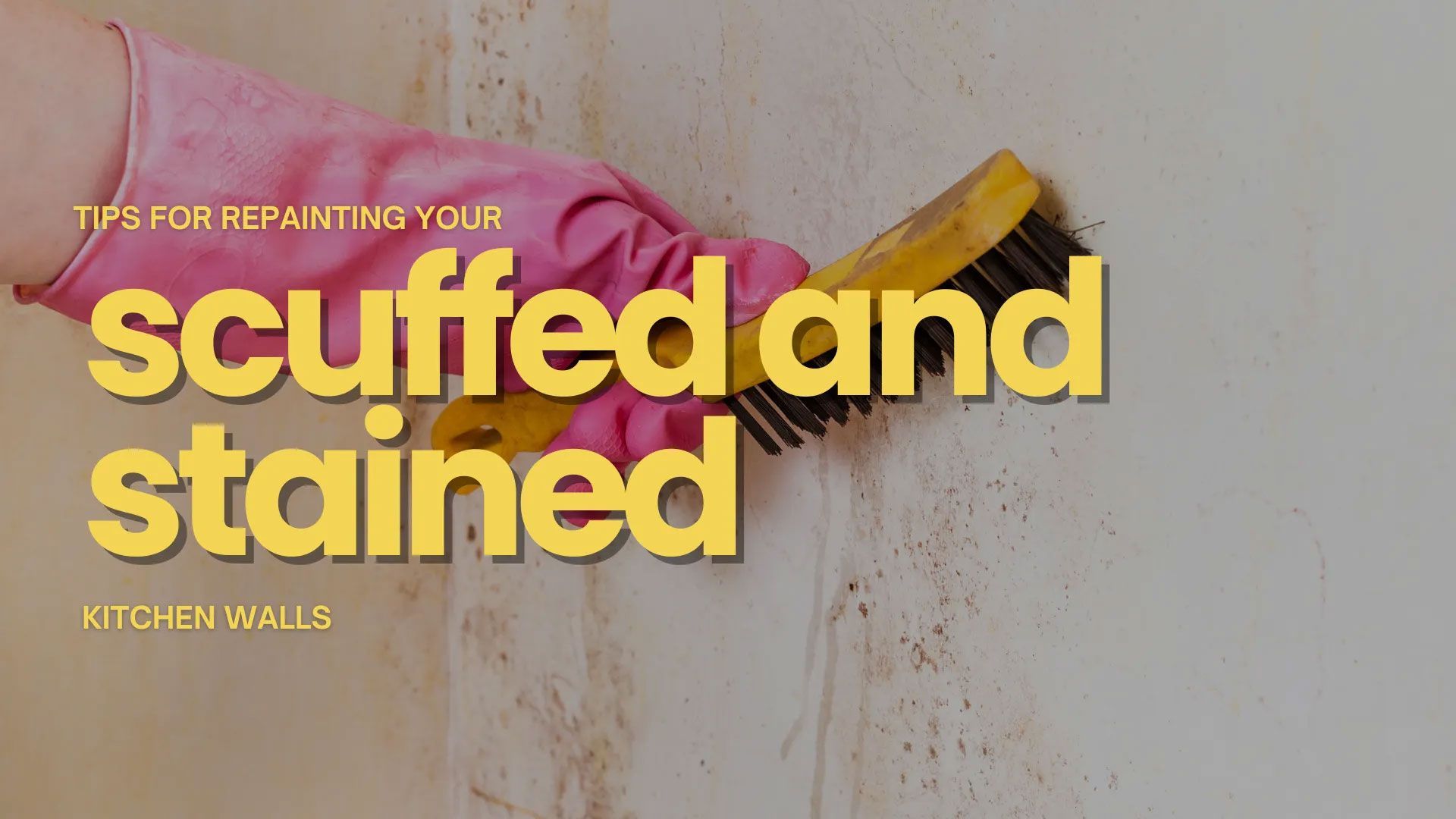 Hand wearing a pink glove scrubbing a scuffed and stained wall with a brush.