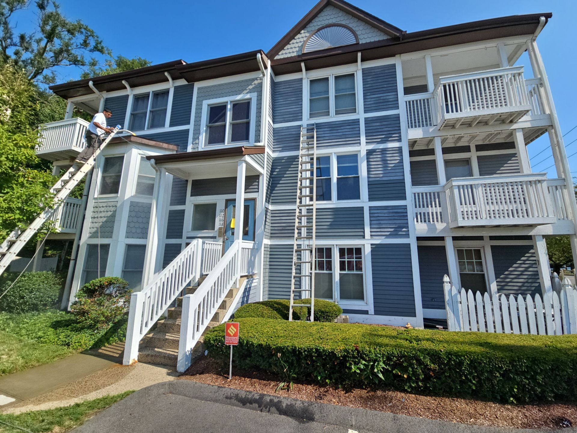 Three-story blue building with white trim; person on ladder; ladders lean on the structure. Sunny day.