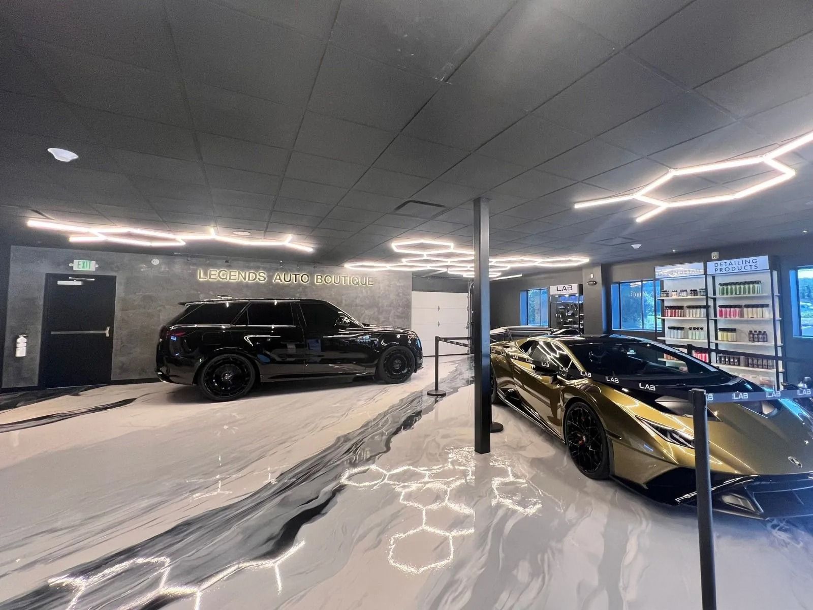 A luxury car showroom featuring a black SUV and a gold Lamborghini. The showroom has unique flooring and hexagonal lighting.