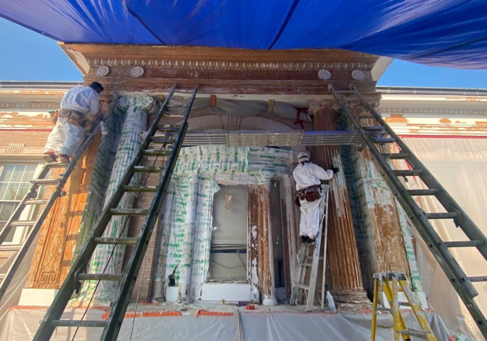 Two workers in protective suits on ladders are restoring a building facade. They are under a blue tarp, with visible damage and scaffolding.