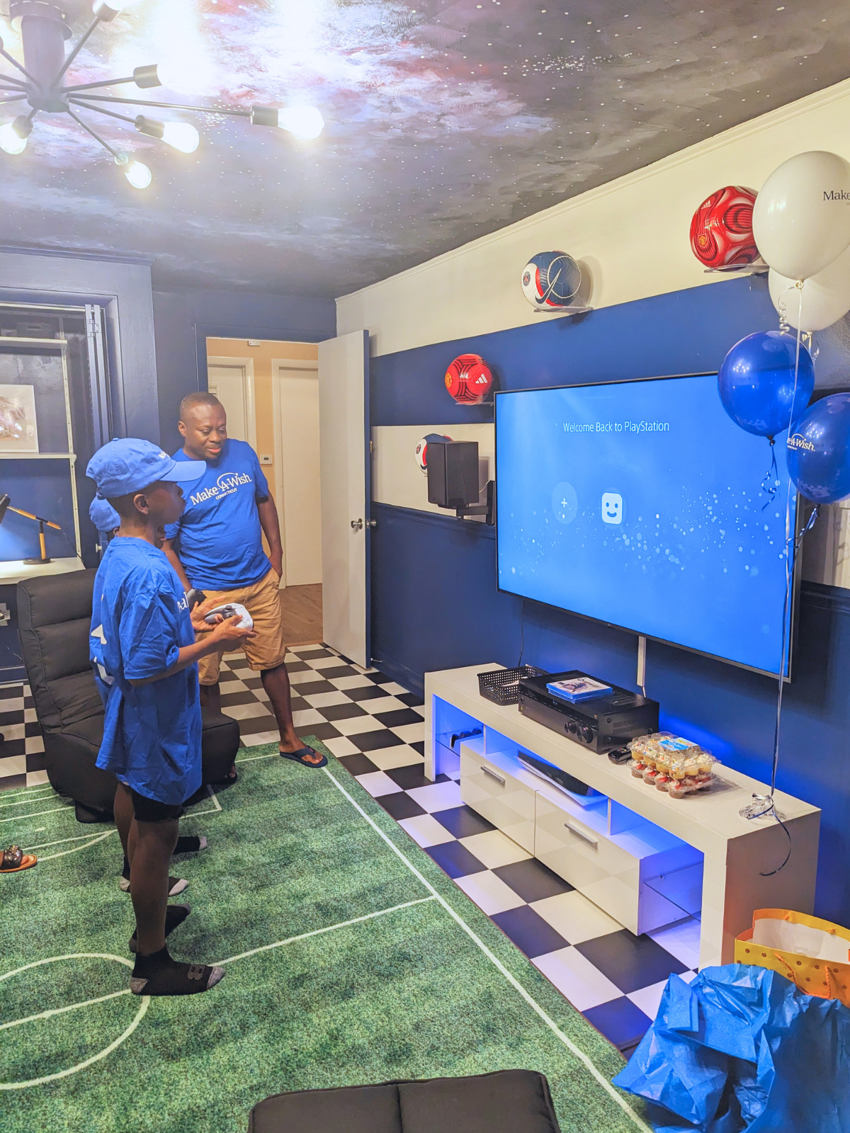 Two people playing video games in a blue and white decorated room with a soccer-themed rug and balloons.