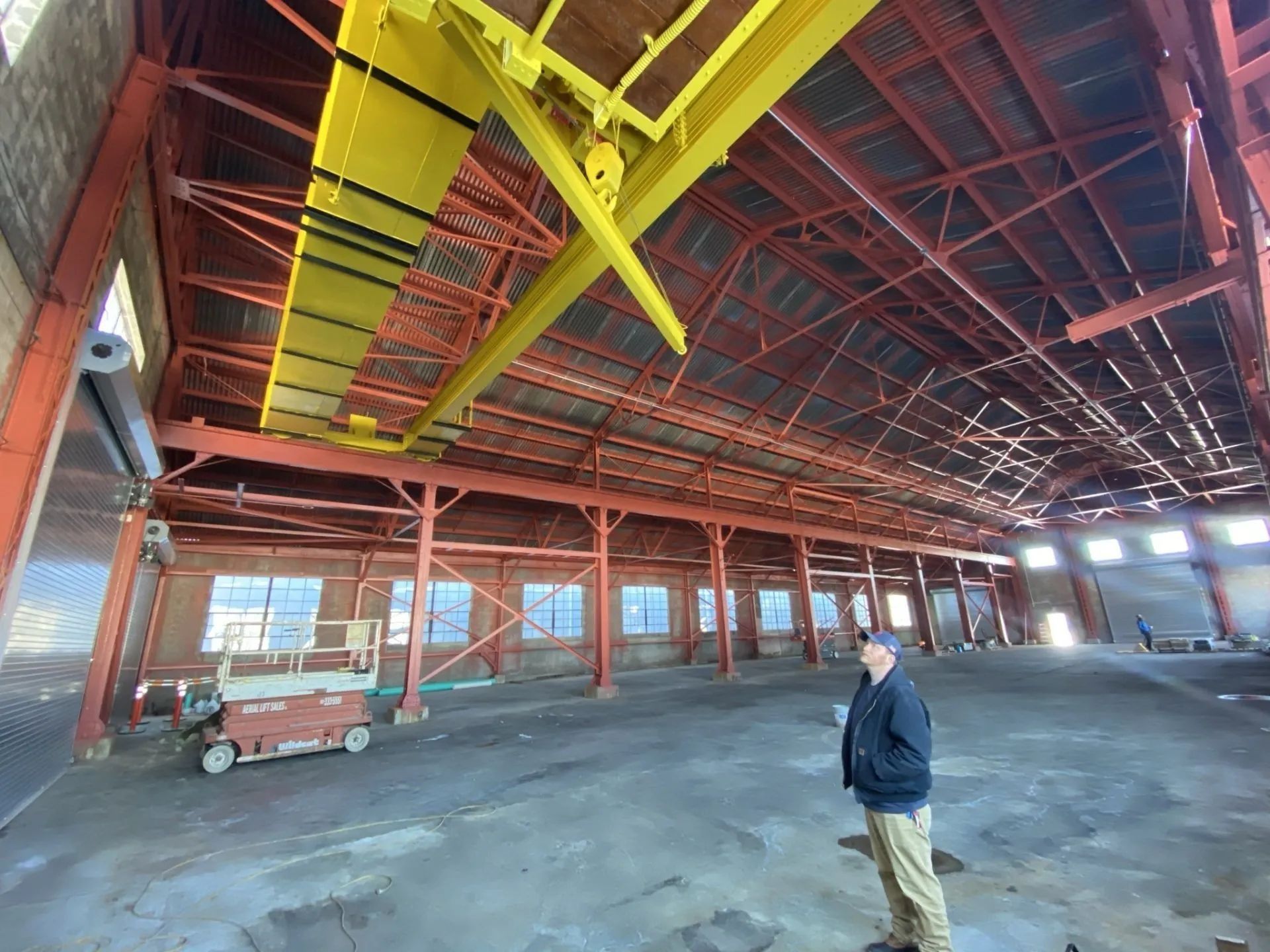 Interior of a large warehouse with red beams, yellow crane, and a man in khaki pants and a dark jacket standing on the concrete floor.