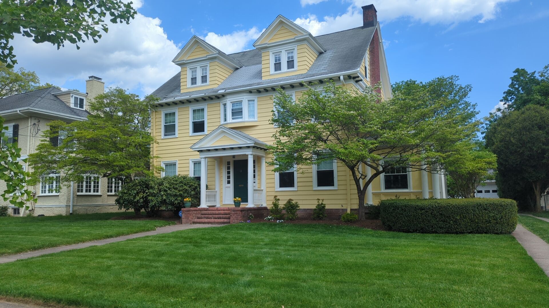 Yellow two-story house with a green lawn under a partly cloudy sky. A tree and bushes are in front.