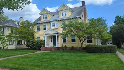 Yellow two-story house with a brick porch, green lawn, and blue sky. A smaller house is partially visible on the left.