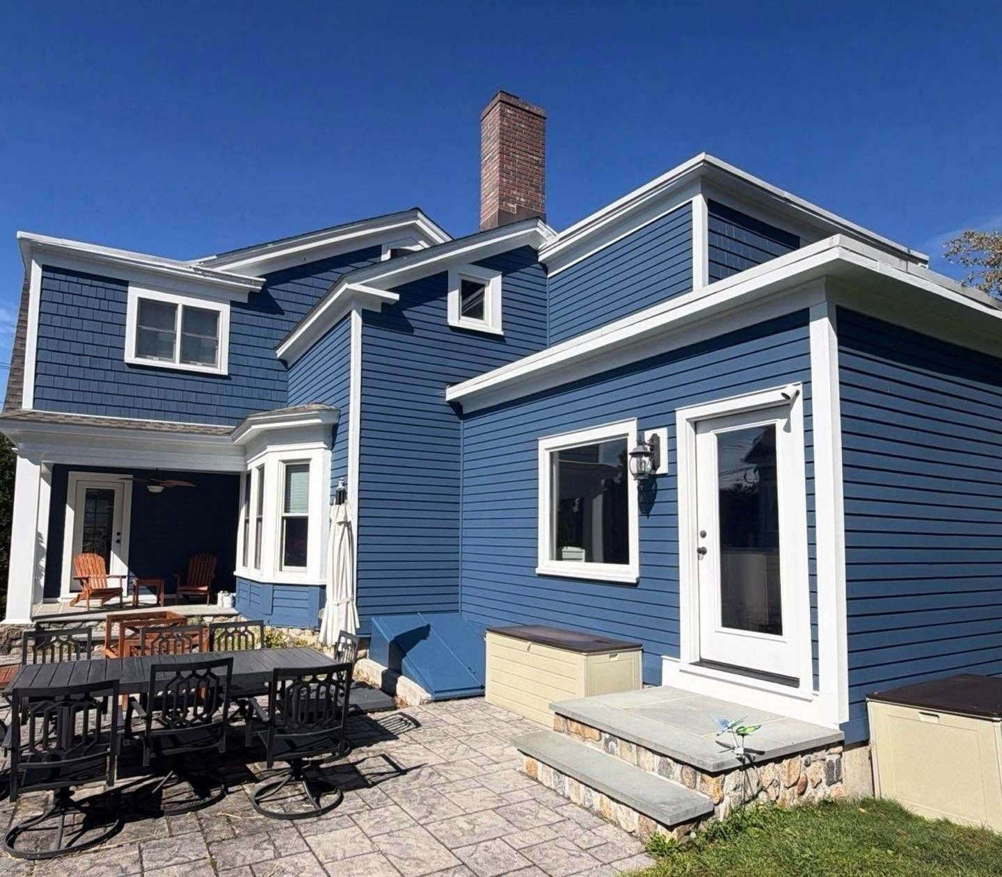 Blue house exterior with white trim, patio furniture, and paved backyard under a clear sky