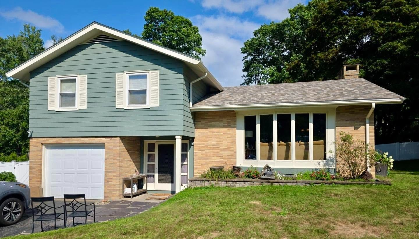 Two-story suburban house with green siding, tan brick, white garage, and a sunlit front lawn