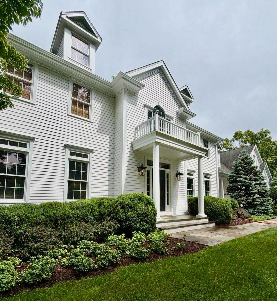 White colonial-style house with columns, balcony, manicured hedges, and green lawn under a cloudy sky