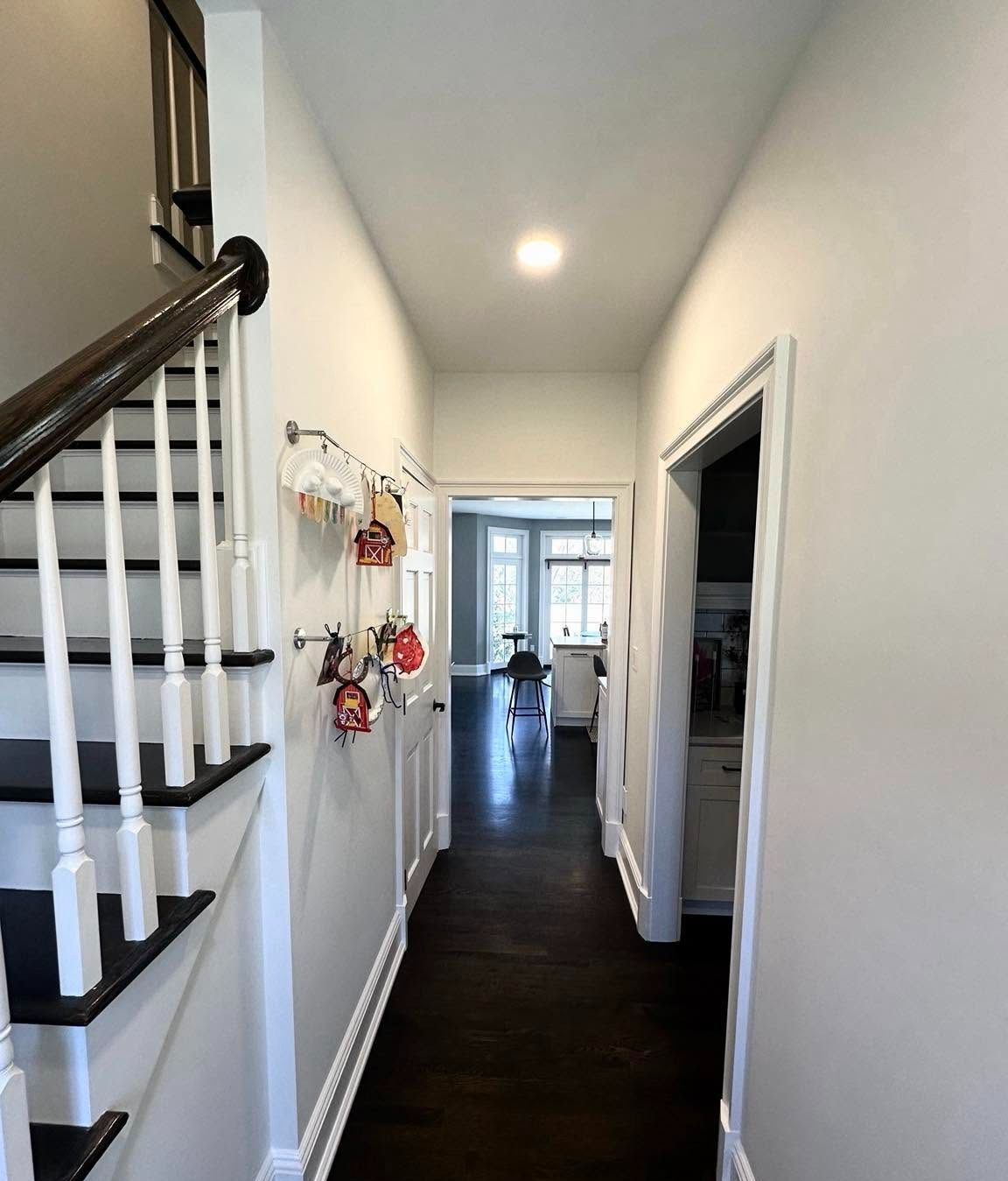Narrow hallway with dark wood floor, white walls, staircase, and open doorway to a bright room.