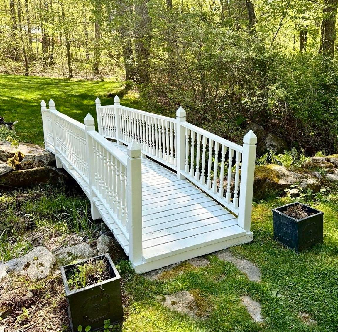 White painted footbridge over a small stream in a wooded garden with green grass and trees