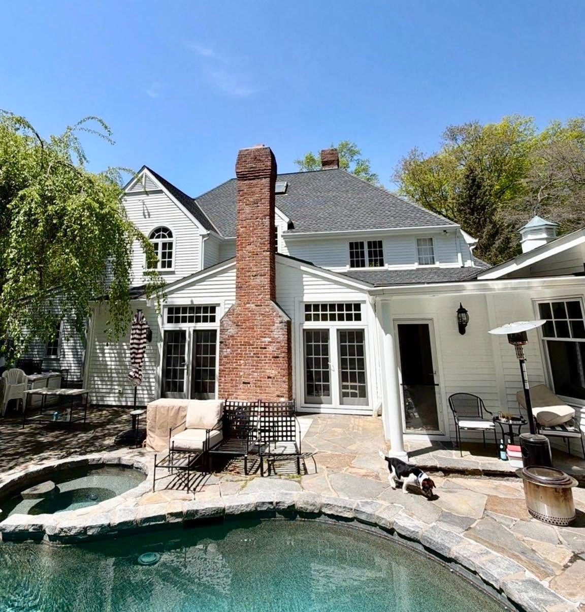 Backyard pool with brick chimney, patio seating, and a white two-story house under a blue sky