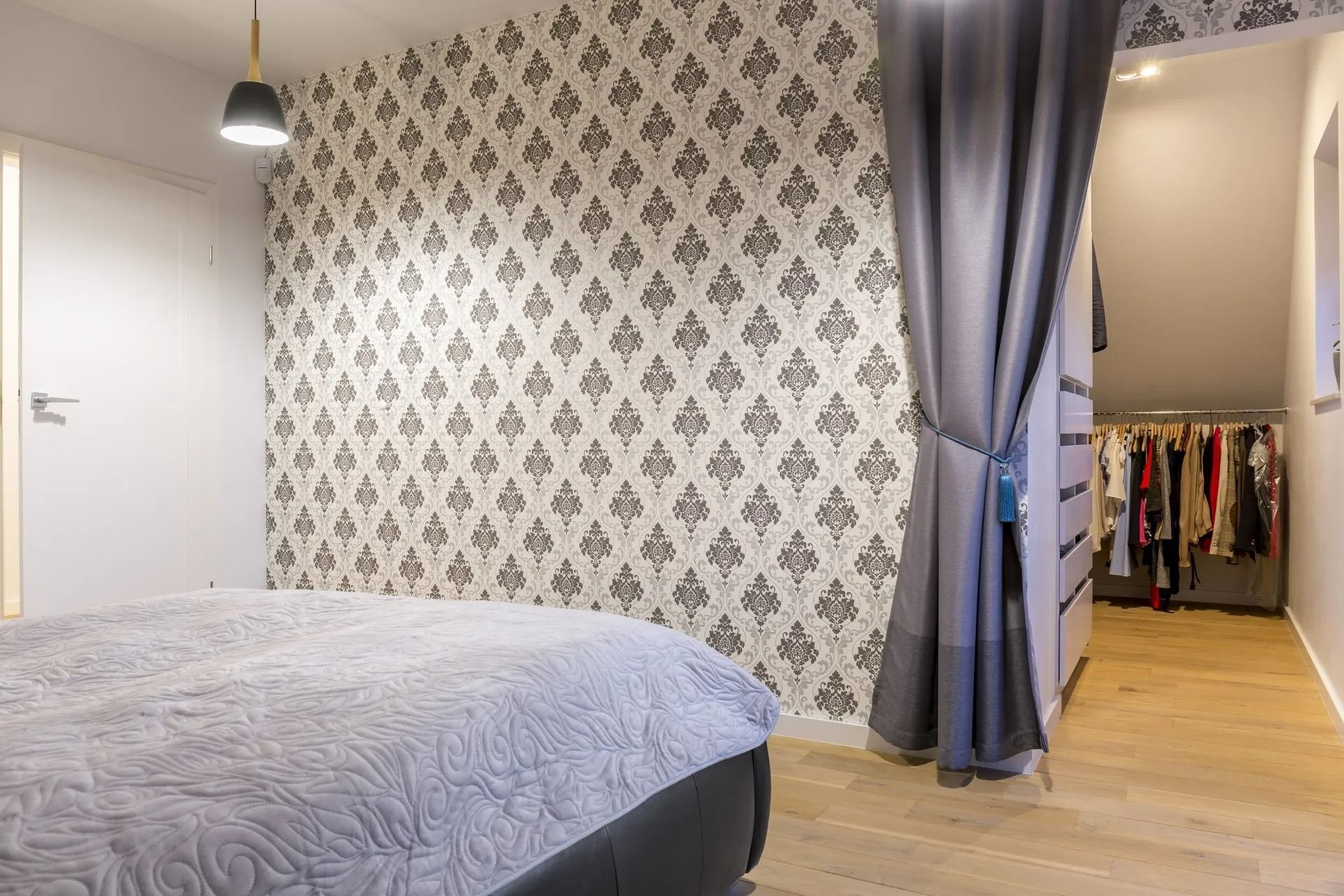 Bedroom with floral wallpaper, a bed, and a closet partially concealed by curtains. Light wooden floors and a hanging light are visible.
