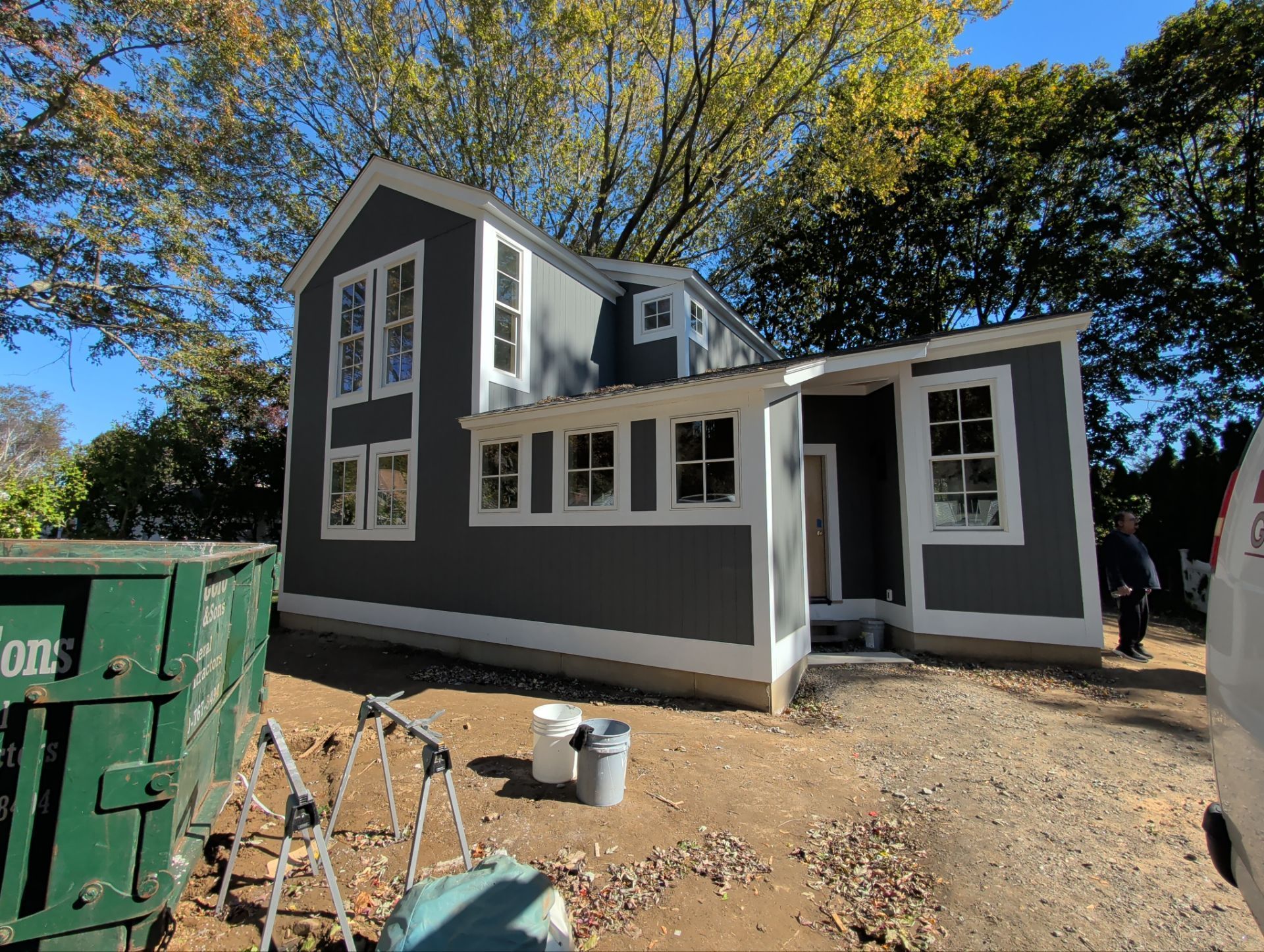 Newly built gray house with white trim, green dumpster, and person in the yard on a sunny day.