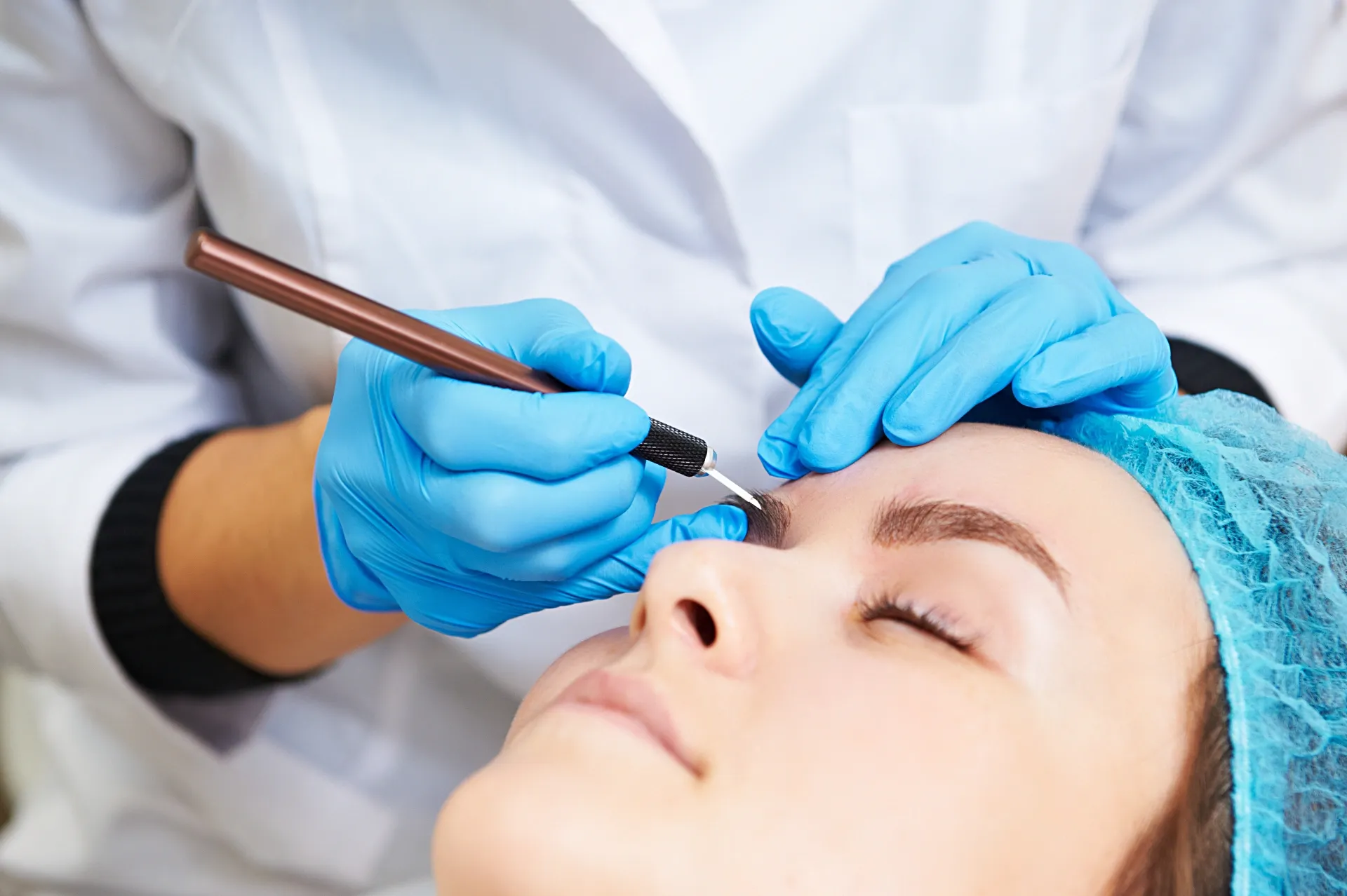 A Person Receiving an Eyebrow Microblading Treatment. A Technician Wearing Gloves Holds a Tool Over a Closed Eye — On Point Cosmetic Tattoo Studio In Noosa, QLD