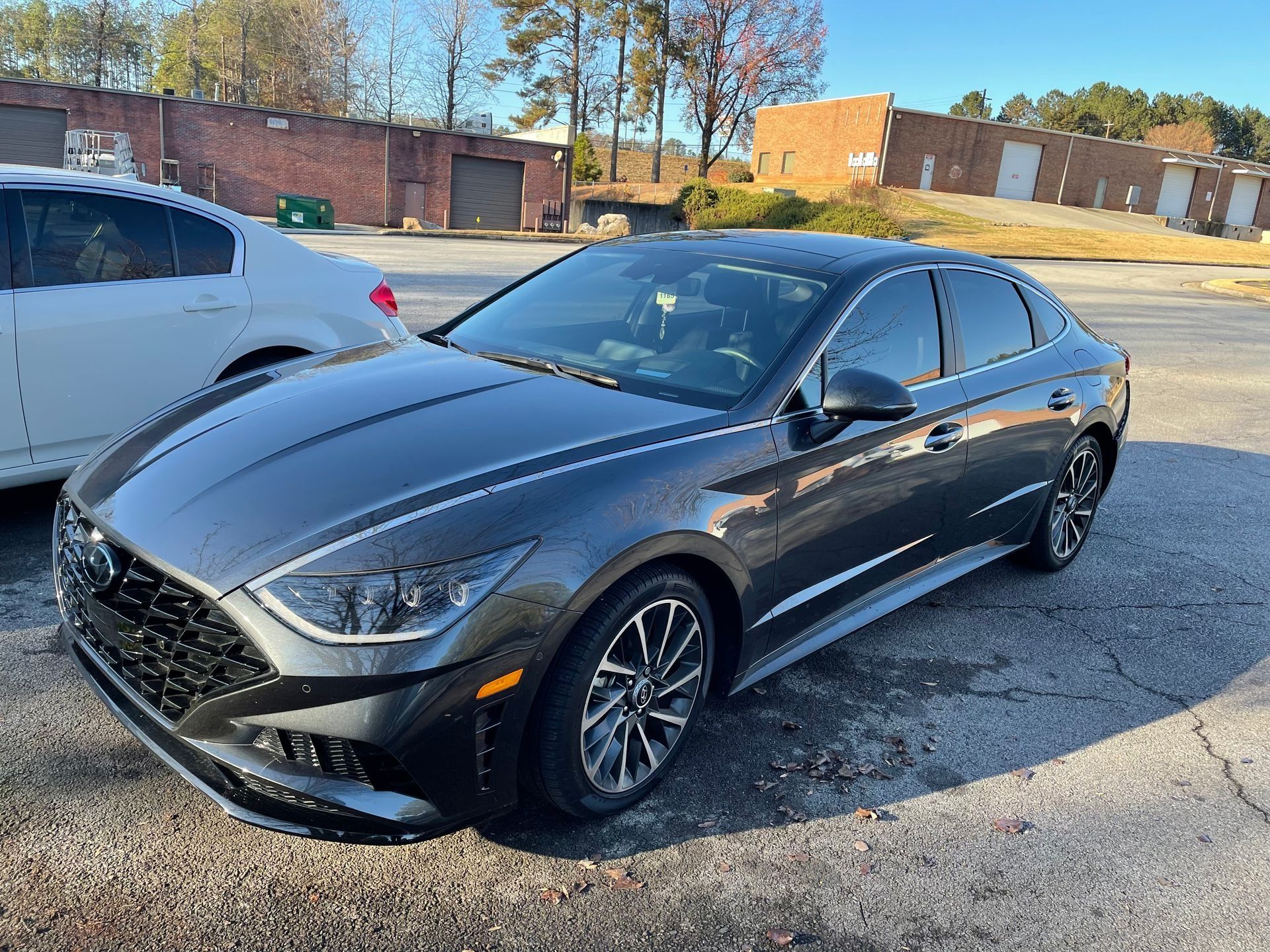 Black sedan parked in a lot beside a white car, with brick buildings and trees in the background