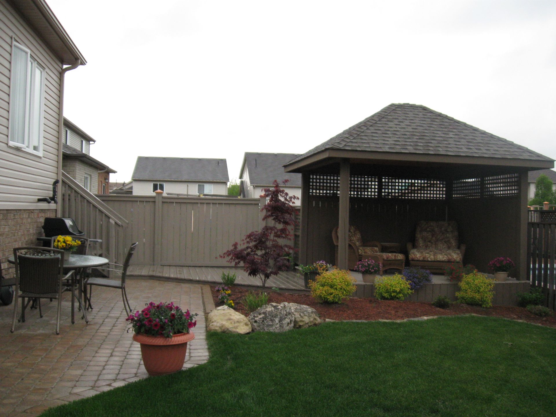 A paved backyard patio with a table, chairs, a potted plant, and a wooden gazebo with outdoor furniture on a grassy lawn.
