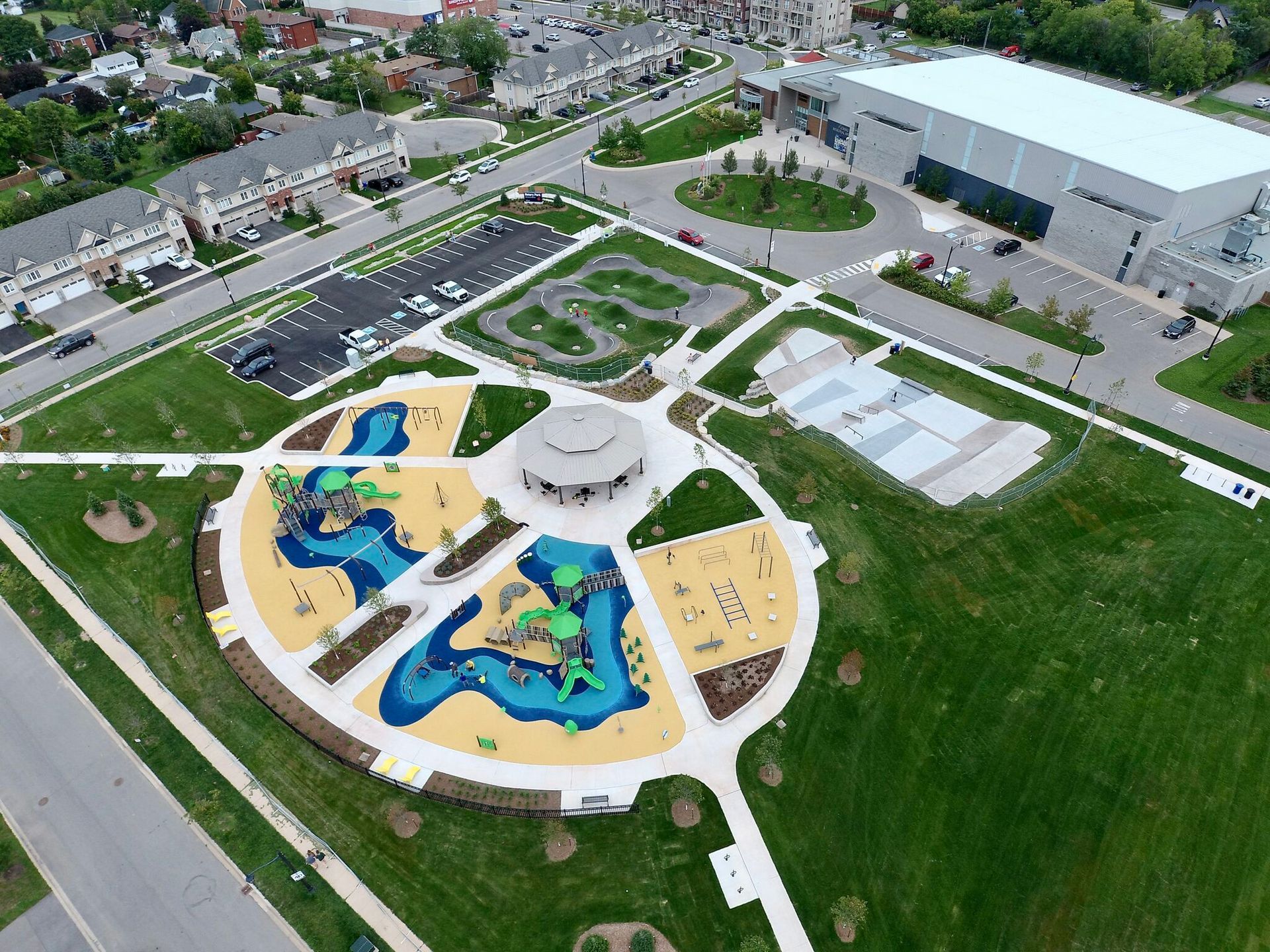 Aerial view of a public park with a circular playground, walking paths, a skate park, and a large building nearby.
