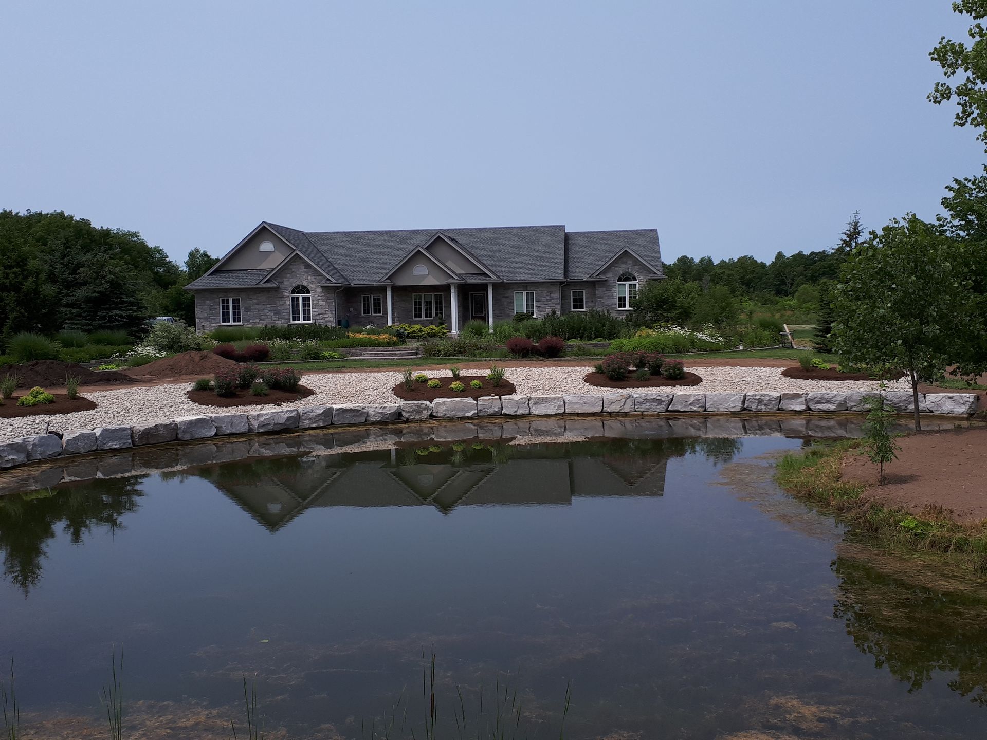 A single-story gray house with a stone facade sits behind a calm pond and a landscaped rock garden.