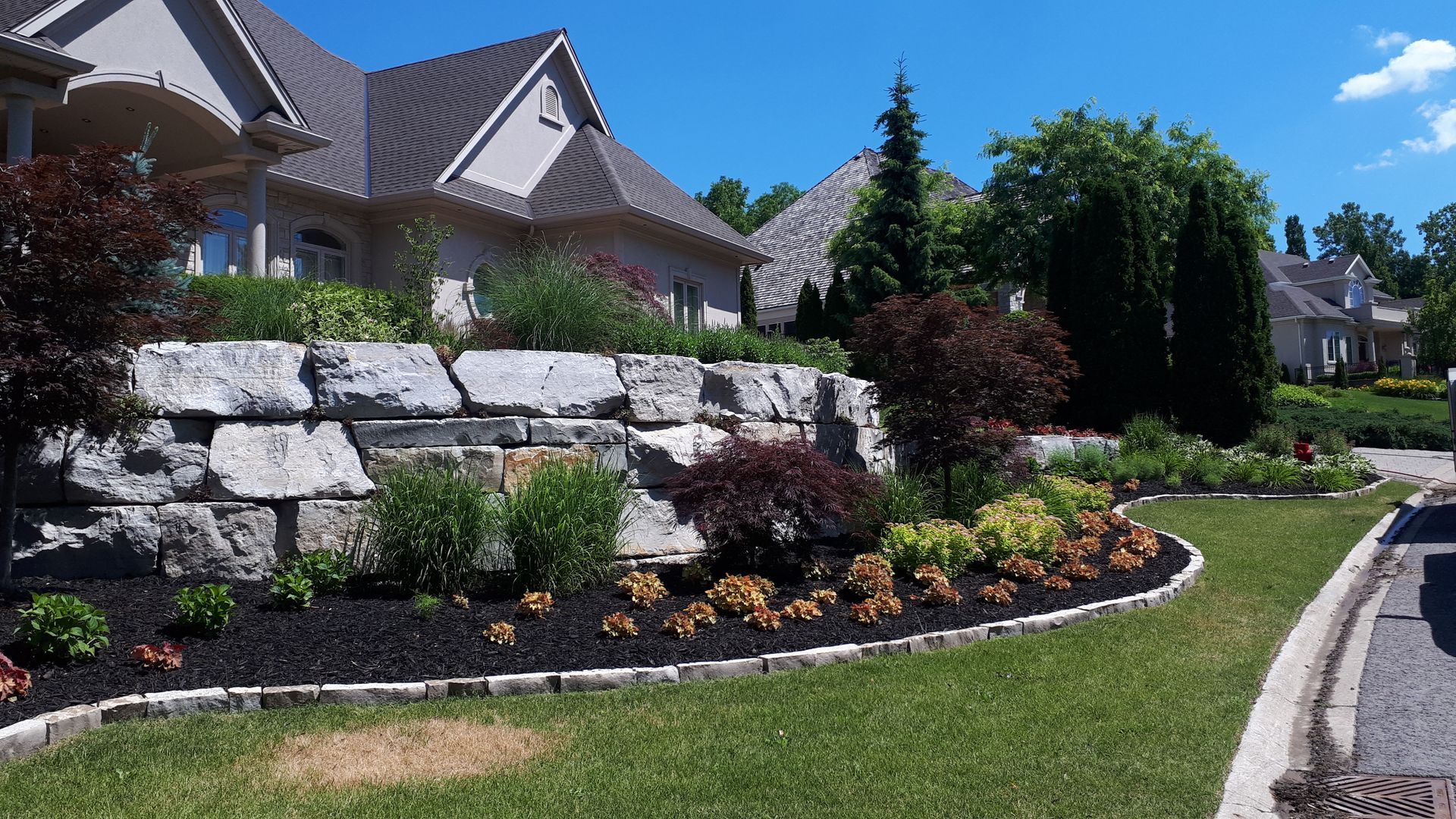 Large stone retaining wall in front of a house, surrounded by landscaped gardens with shrubs and mulch on a sunny day.