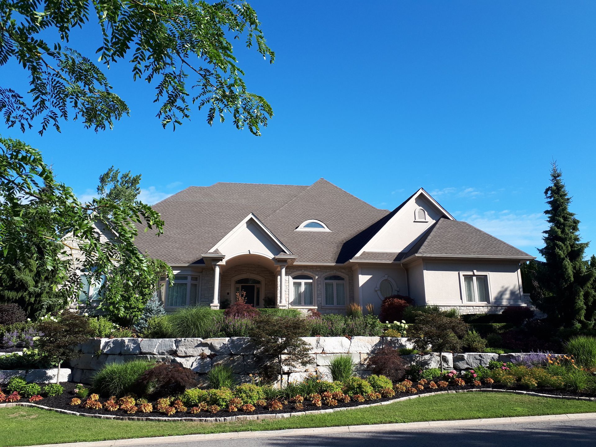 A large, light-colored house with a dark roof sits behind a tiered stone retaining wall and a manicured garden.