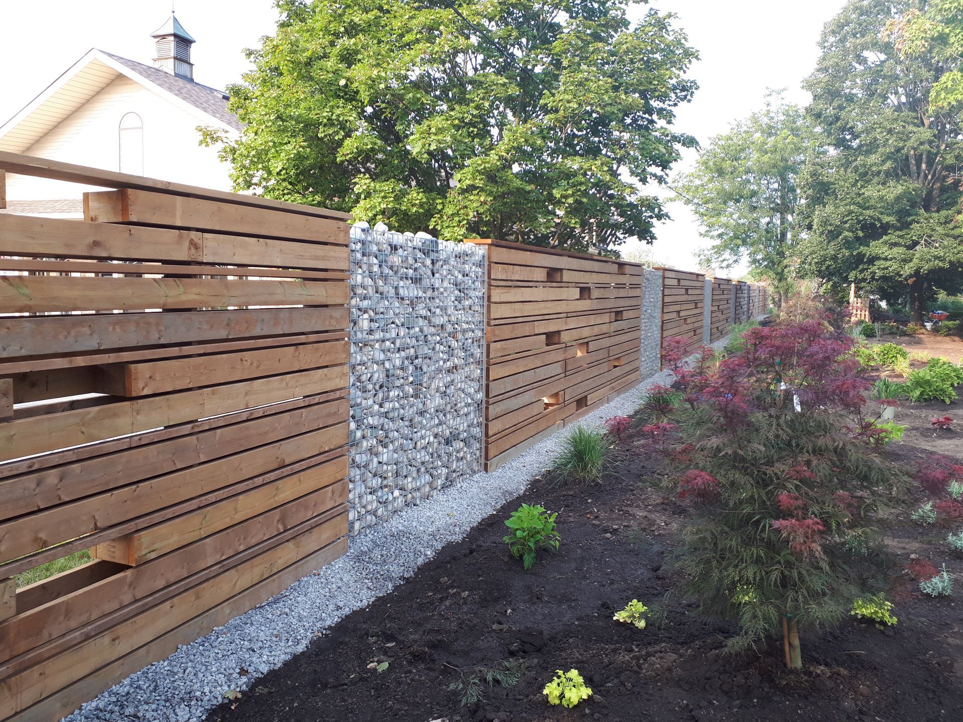 A modern outdoor fence alternating horizontal wood slat panels and vertical columns of stacked grey stones in a garden.