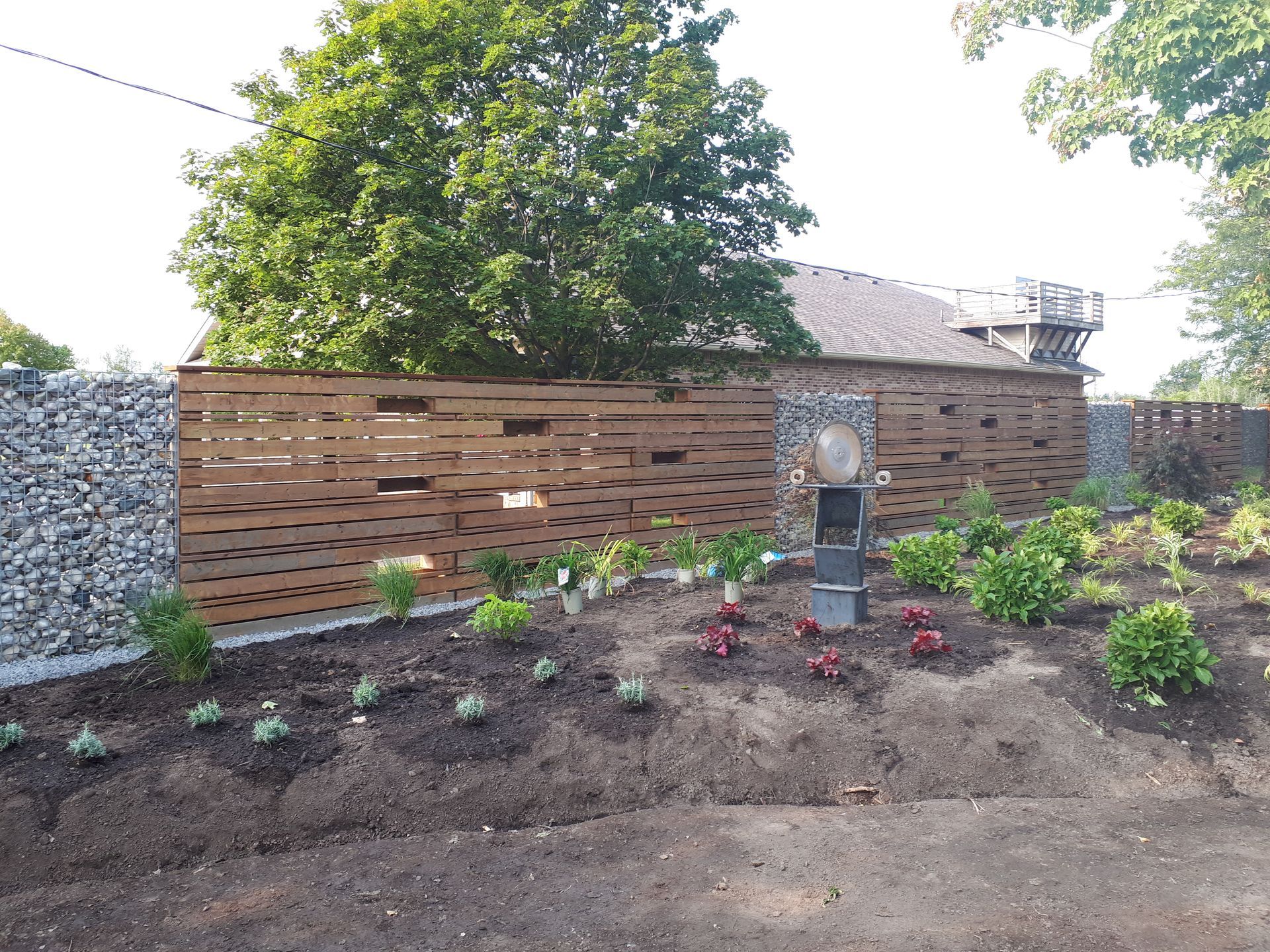 A wooden fence with open gaps, flanked by two stone gabion pillars, set behind a newly mulched garden bed.