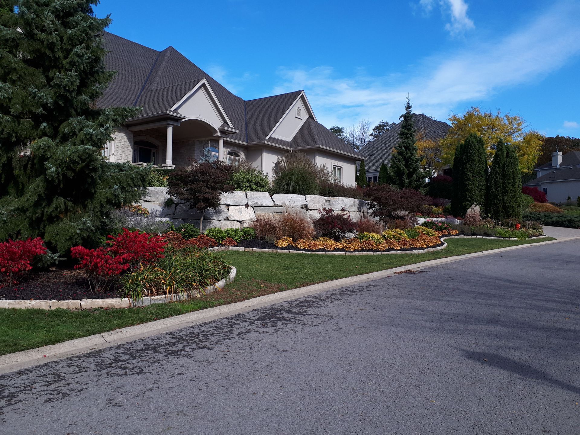 A large suburban house with a stone retaining wall, landscaped gardens, and tall evergreens under a blue sky.