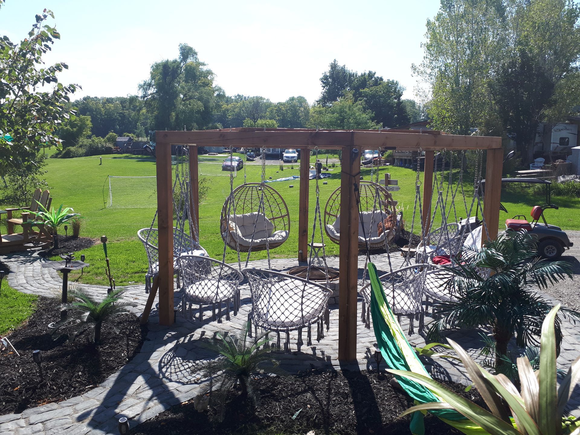 A wooden pergola structure stands in a garden, framing several white hanging egg chairs set on a paved stone patio.
