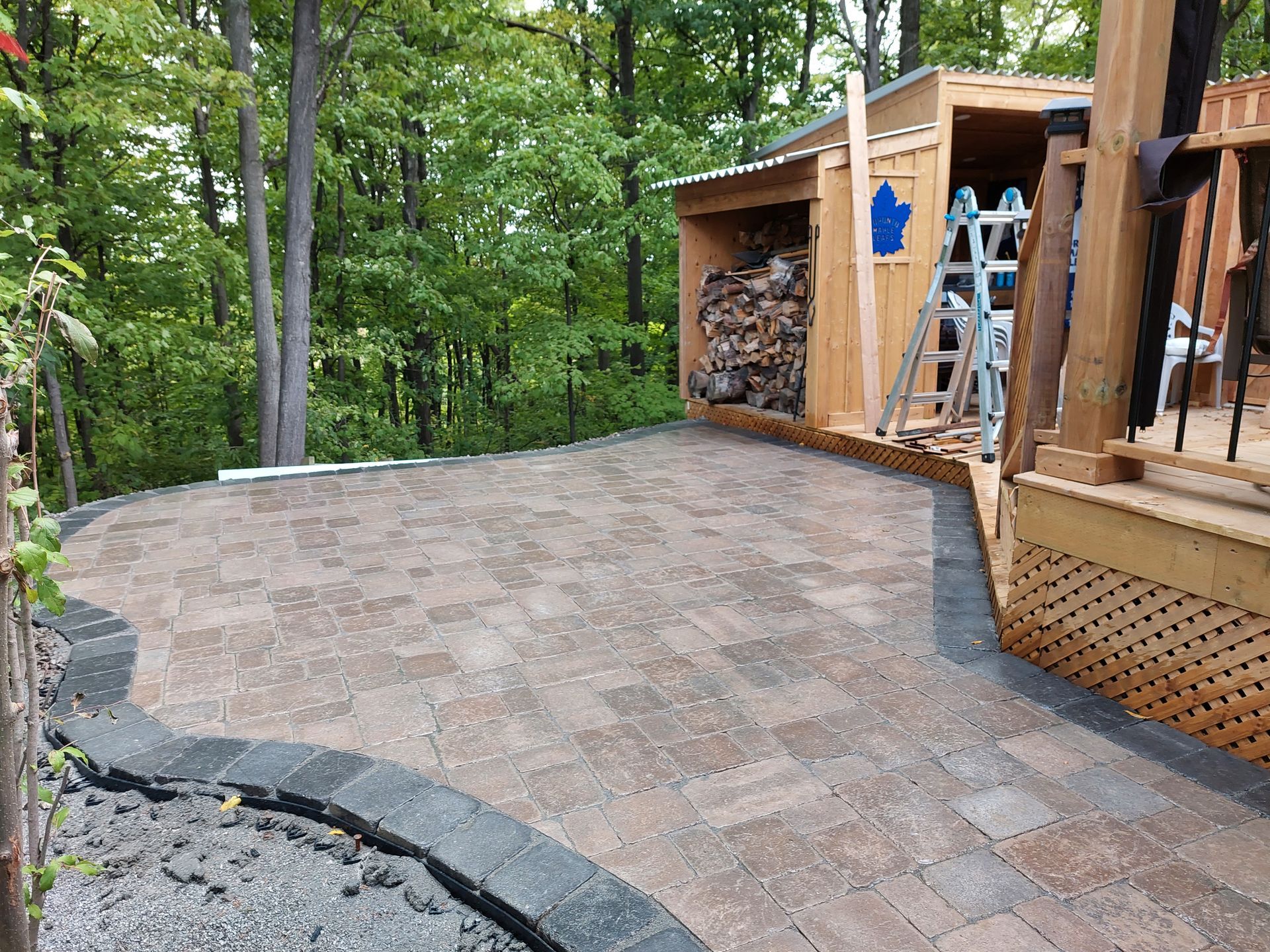 A patio of earth-toned pavers with a dark border, set against a wooden shed, a deck, and a wooded background.