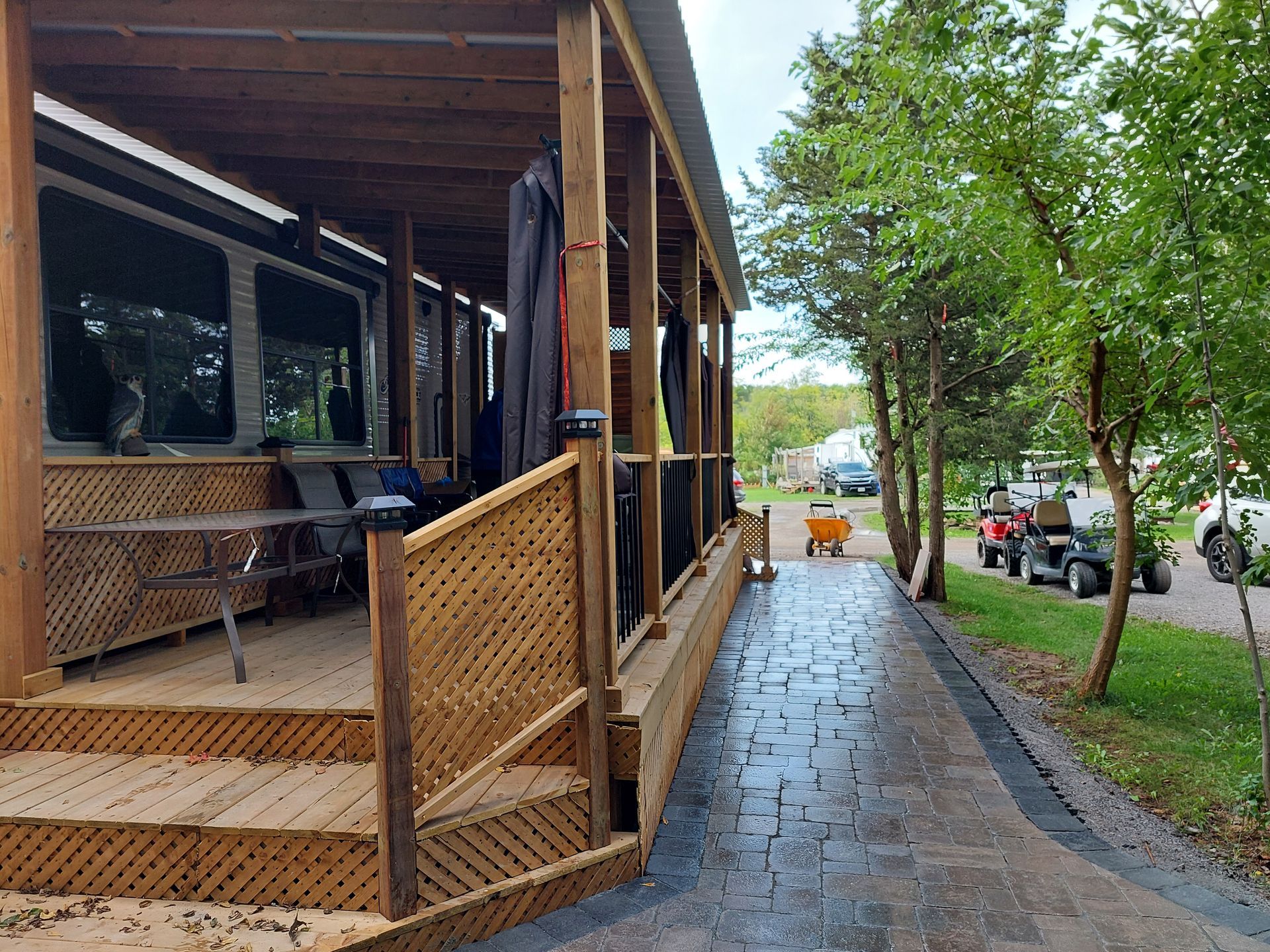 A wooden deck with a covered patio alongside a stone walkway, with golf carts parked in the grass area nearby.