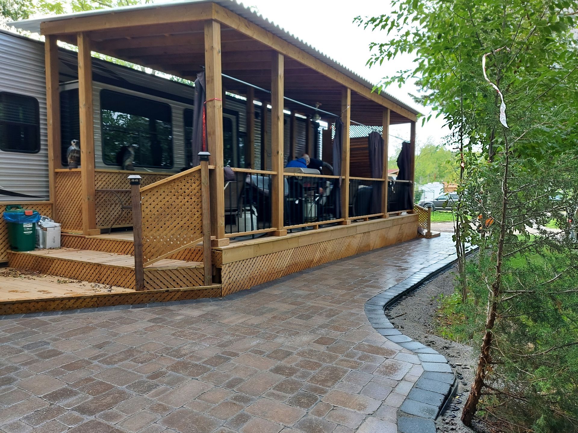 A wooden deck with a covered roof attached to an RV, set beside a paved stone patio in a wooded area.