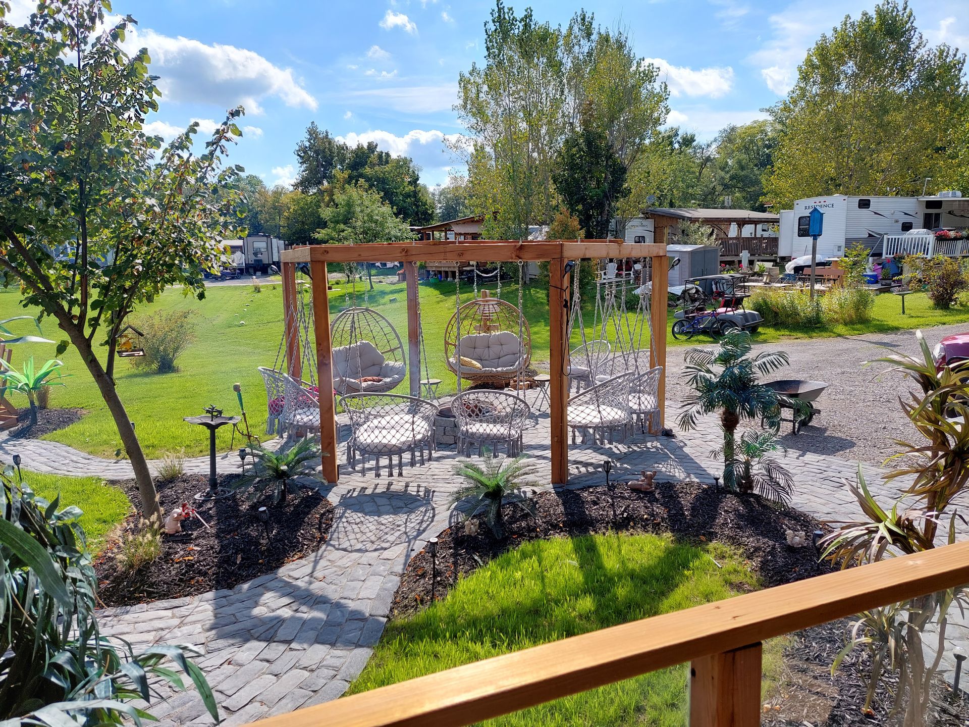 A wooden pergola with hanging chairs sits on a gravel patio in a grassy campground with trees and RVs in the background.