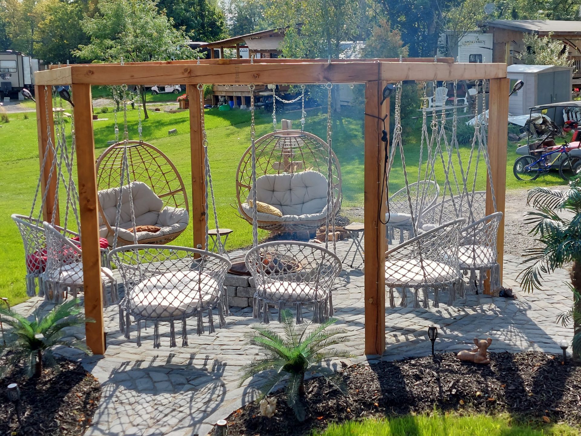 An outdoor pergola area with wicker-style hanging chairs and seating, set on a stone patio in a grassy yard.