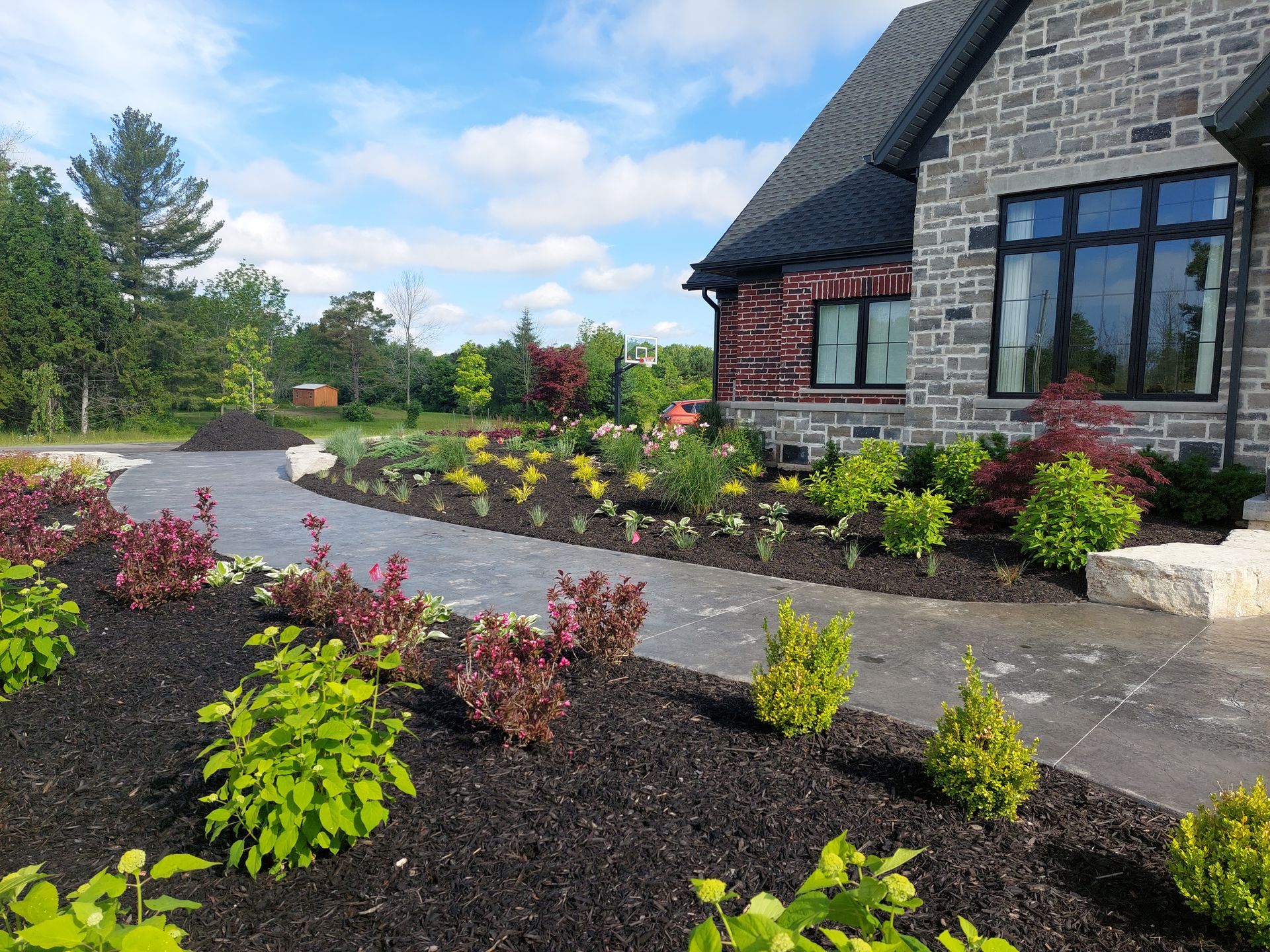 A stone house exterior with a paved walkway surrounded by neatly mulched garden beds featuring colorful shrubs and greenery.