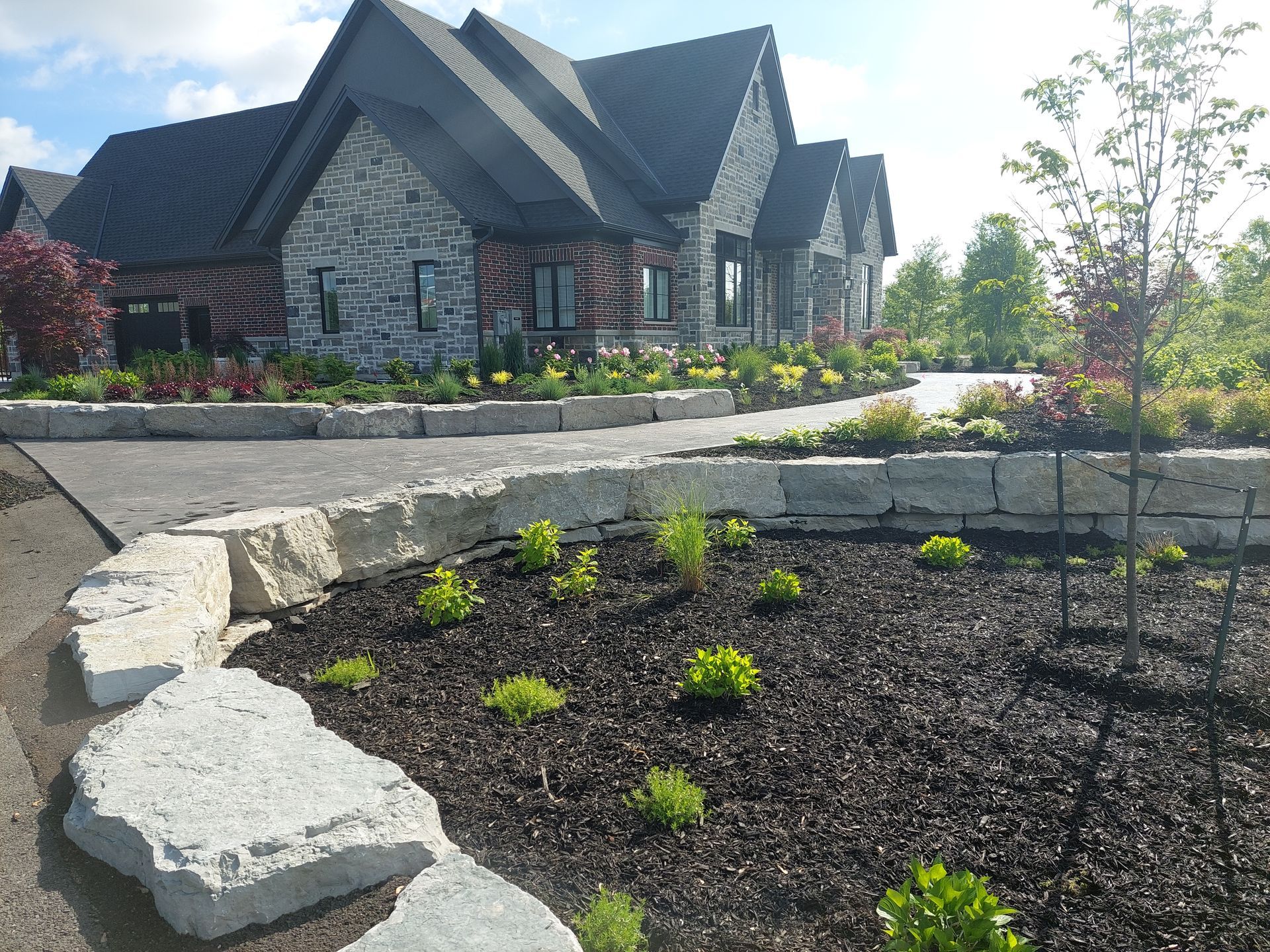 A stone-faced house with dark roofs stands behind a stone retaining wall and a landscaped garden bed with mulch.