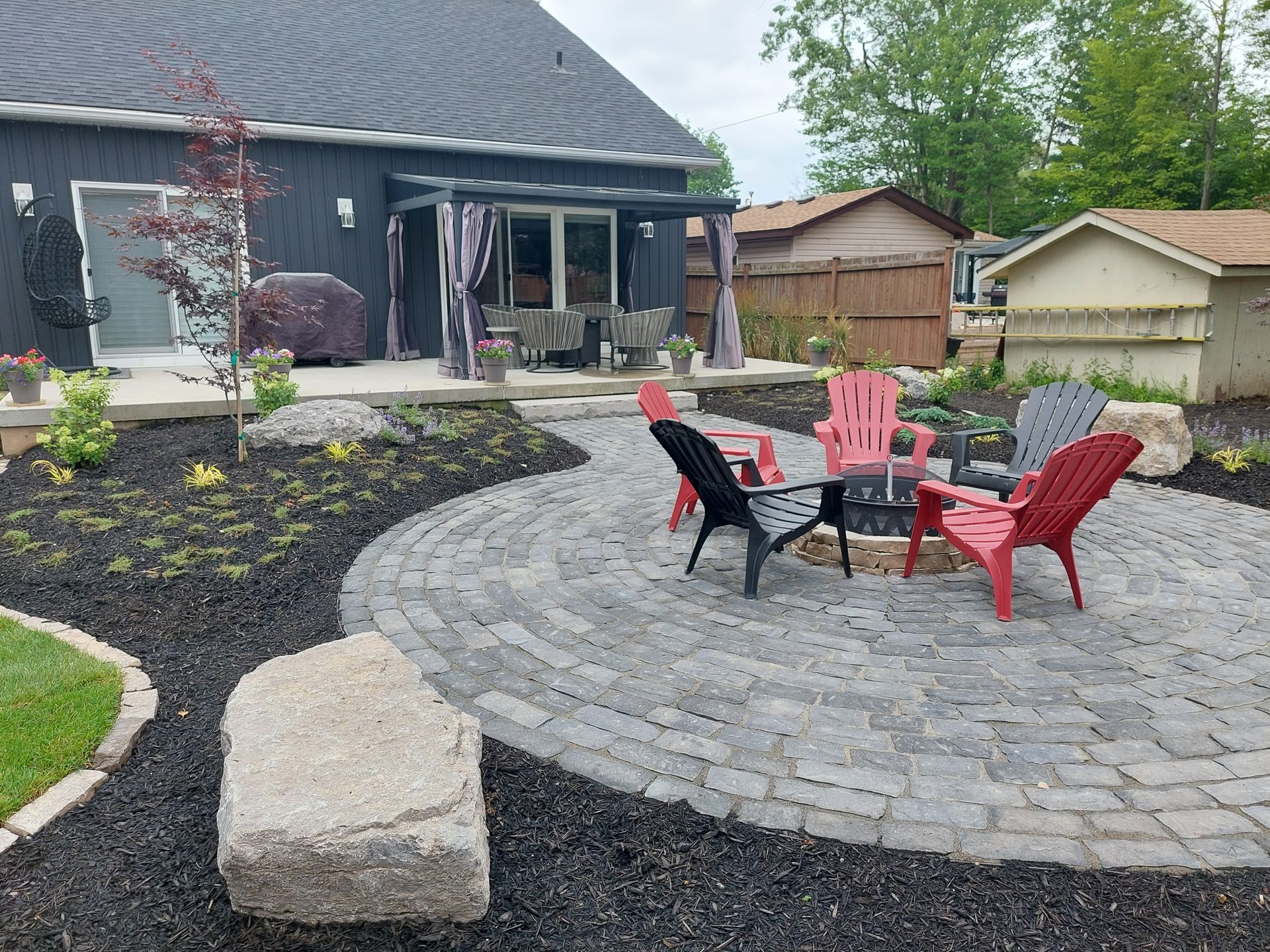 A backyard patio featuring a gray paver circle with red and black chairs, surrounded by dark mulch and large boulders.