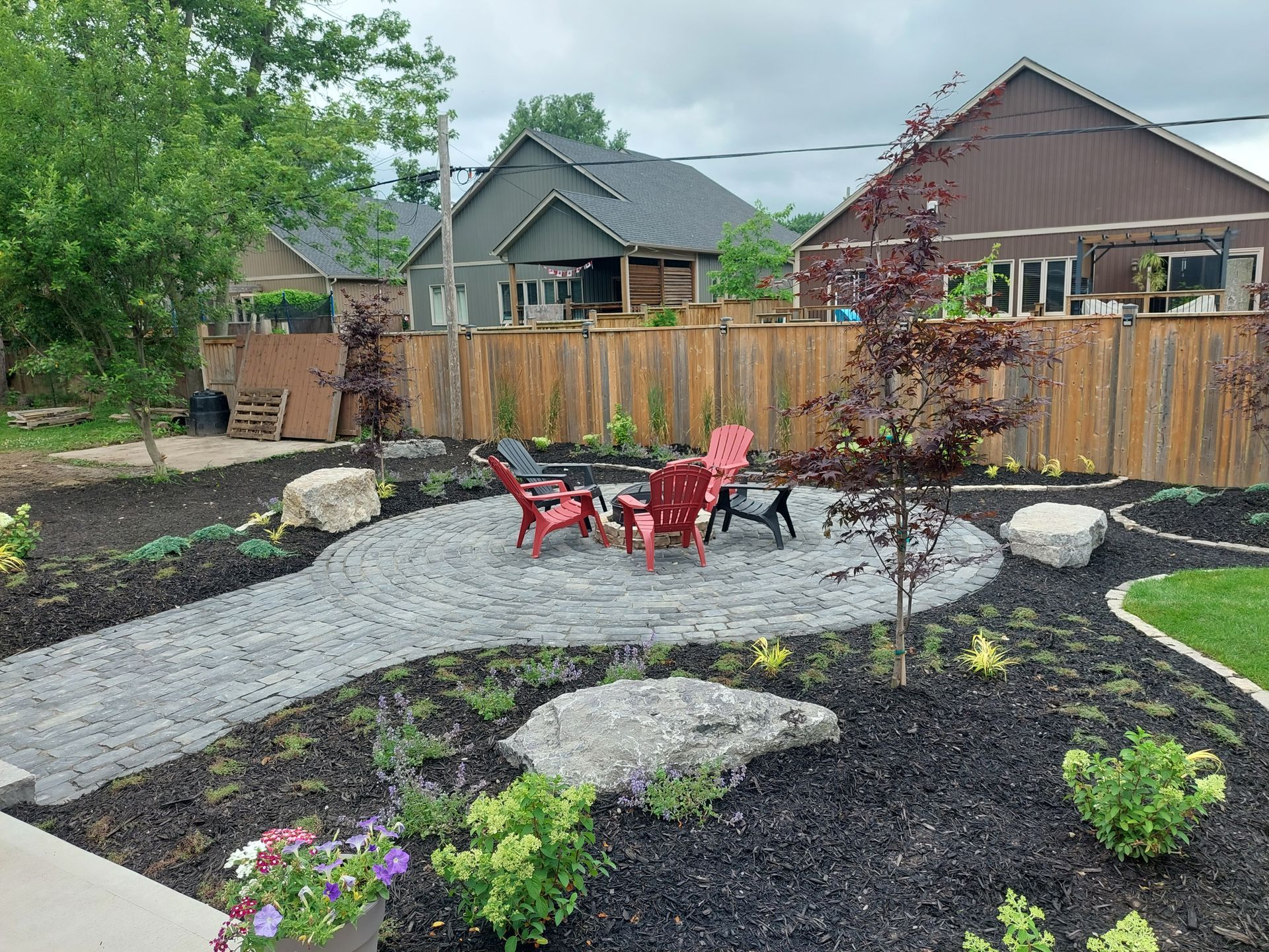 A backyard fire pit area featuring a stone pathway leading to a circular patio with four red chairs, surrounded by mulch.